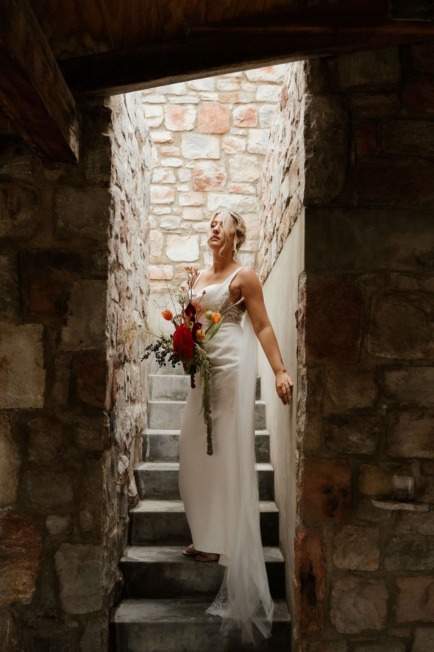 A bride in a flowing white dress stands in a stone archway holding a red bouquet on stone steps.