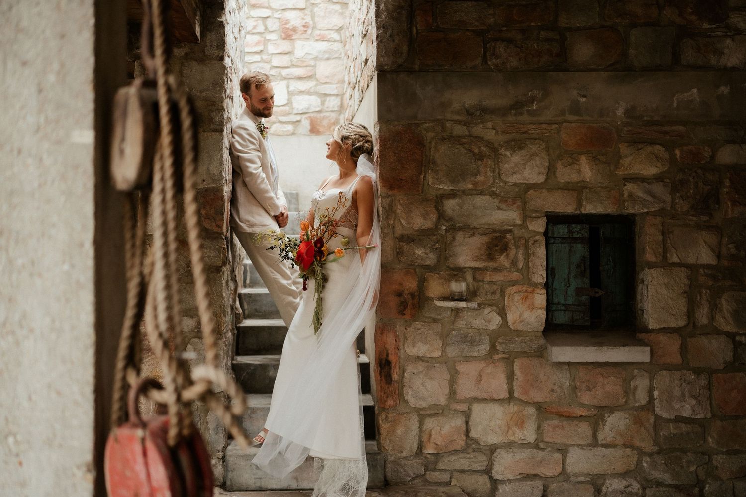 Two people in wedding attire share a moment on vintage stone steps surrounded by rustic architecture.
