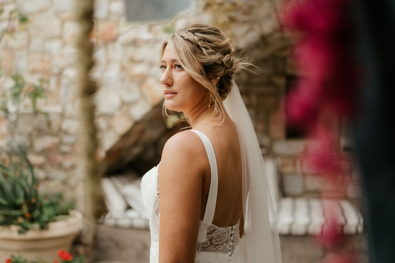 Side profile view of person wearing white gown and veil against stone wall with blooming flowers.