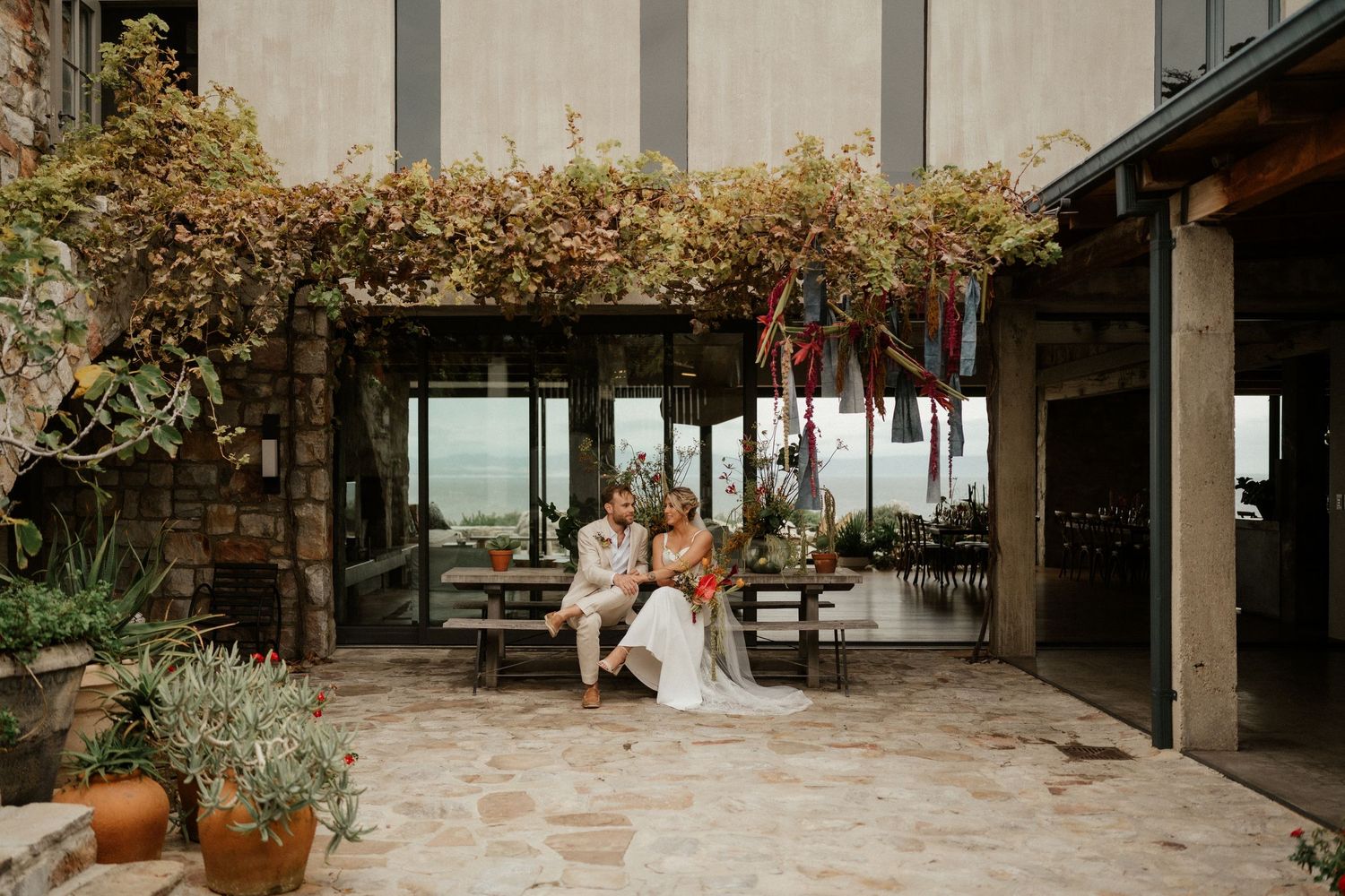 A couple sits beneath a lush vine-covered pergola on a stone patio with mountain views in the background.