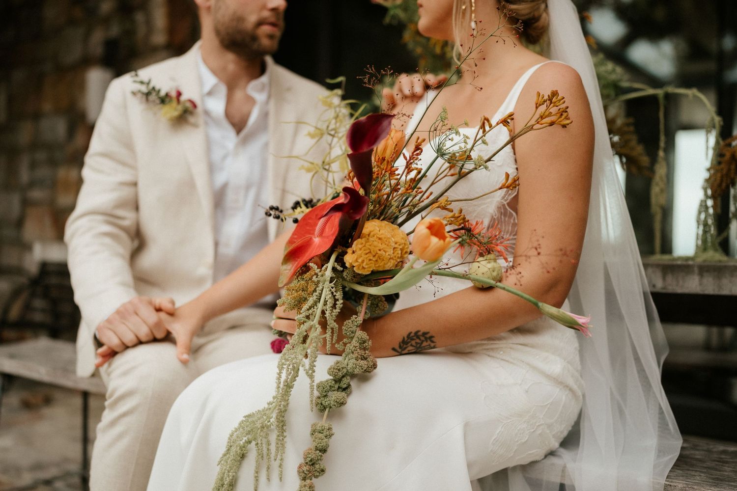 A couple in light colored wedding attire sitting together while holding a vibrant autumn-toned bouquet.