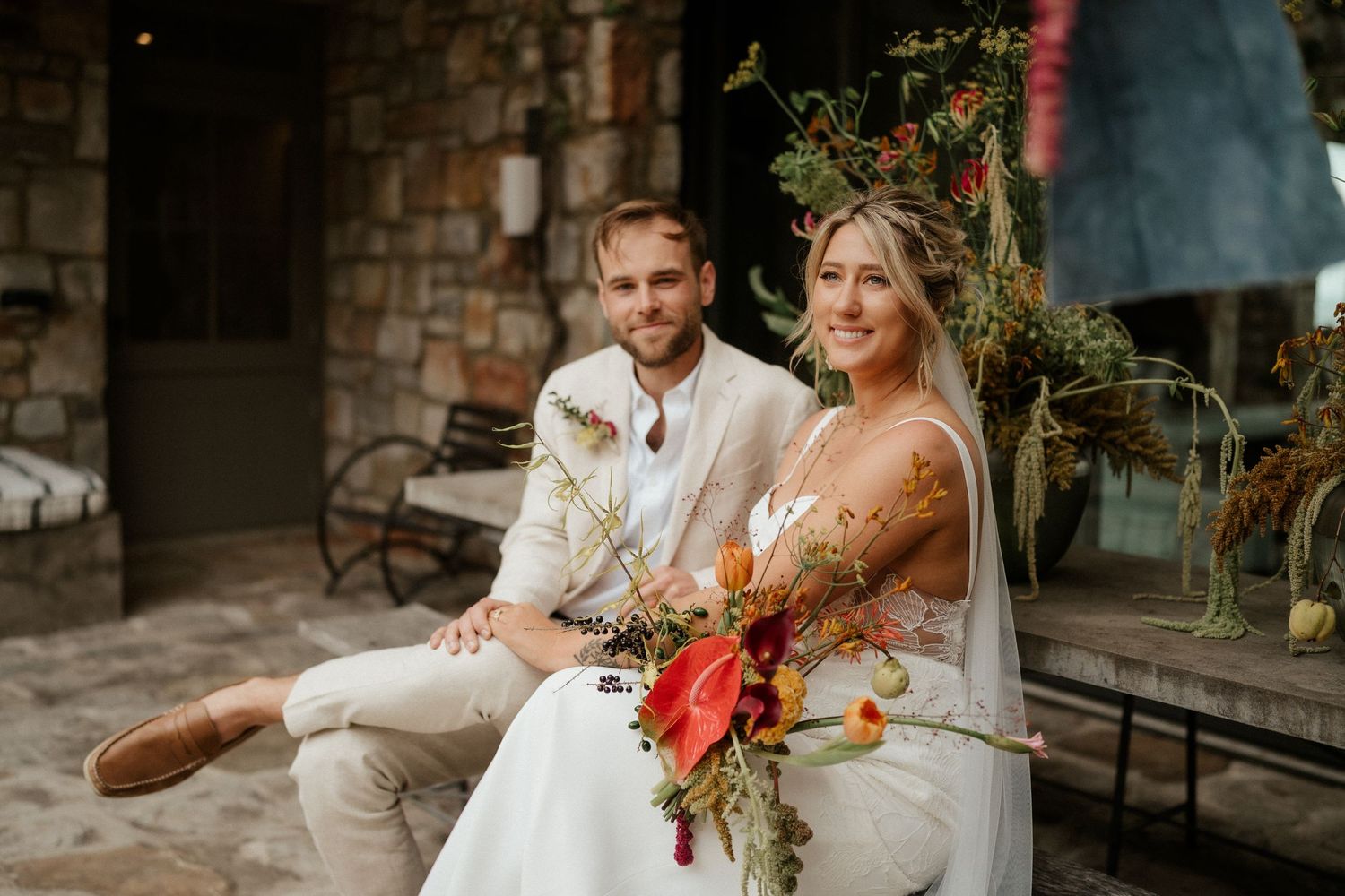 A couple in wedding attire sits with autumn-colored floral arrangements against a stone wall exterior.