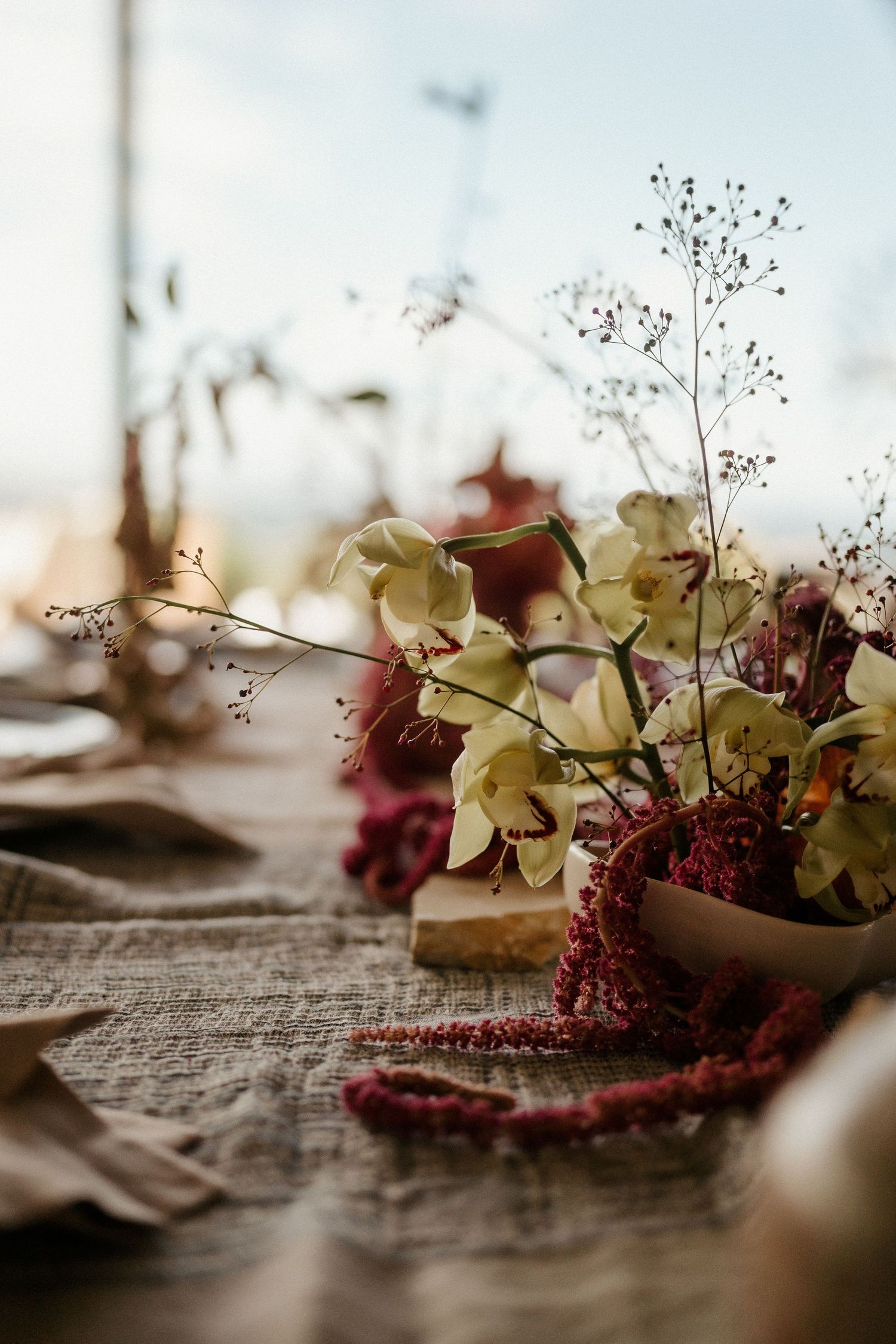 Elegant floral centerpiece with pale yellow flowers and burgundy accents on a rustic textured tablecloth.
