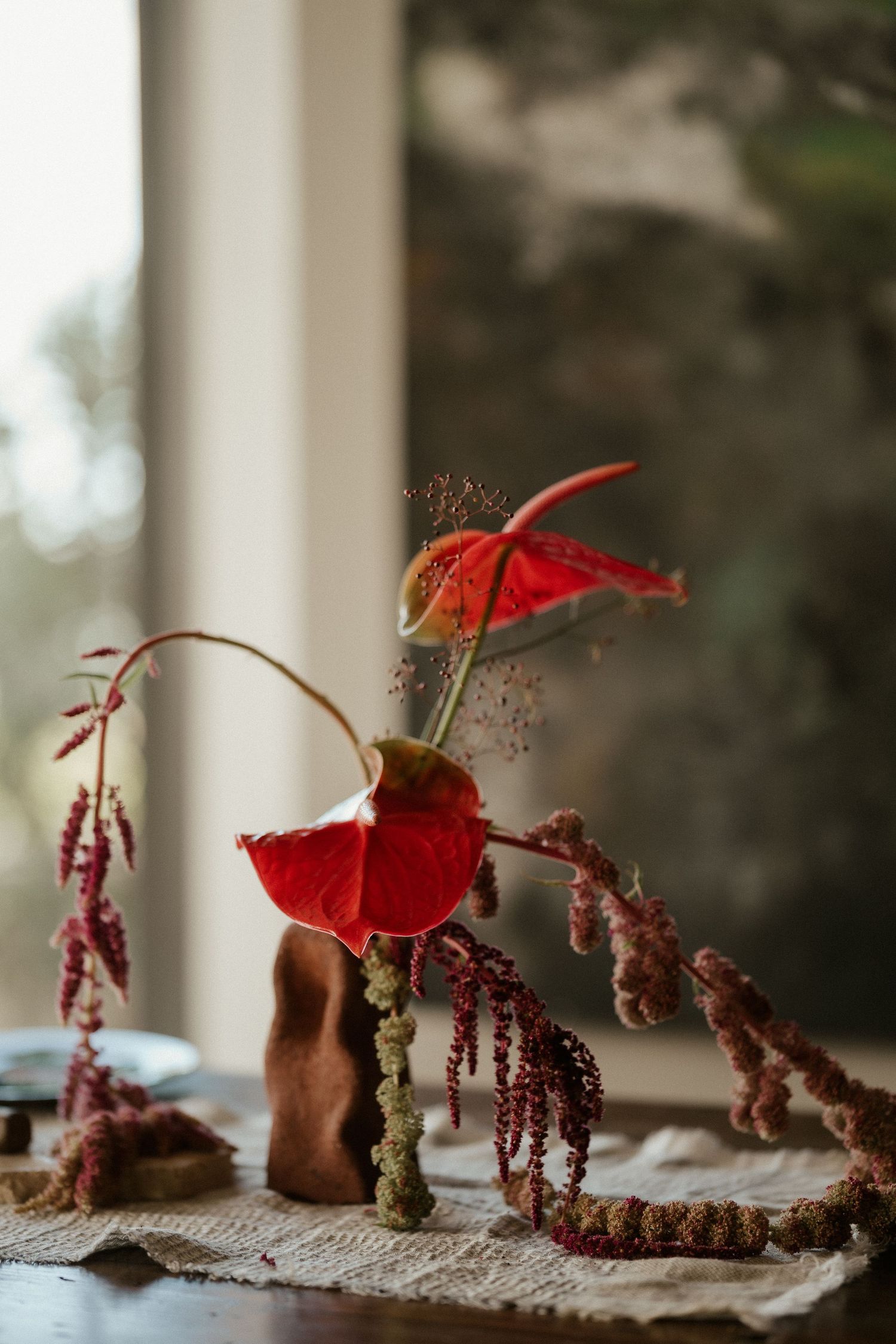 Artistic arrangement of red anthurium flowers and dried botanicals on a rustic windowsill with natural lighting.