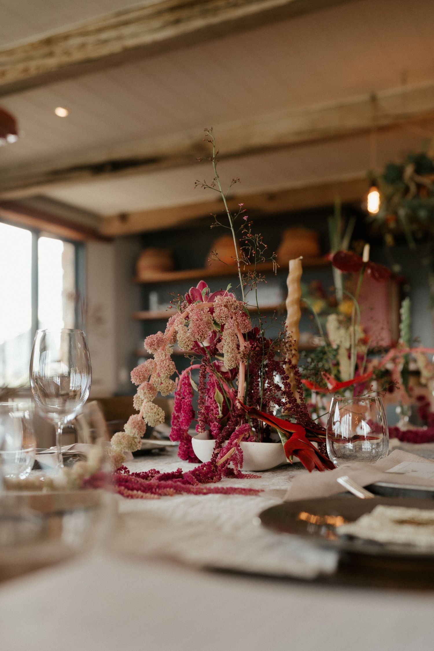 Elegant wedding table setting with pink and red floral centerpieces and wine glasses in a rustic indoor venue.