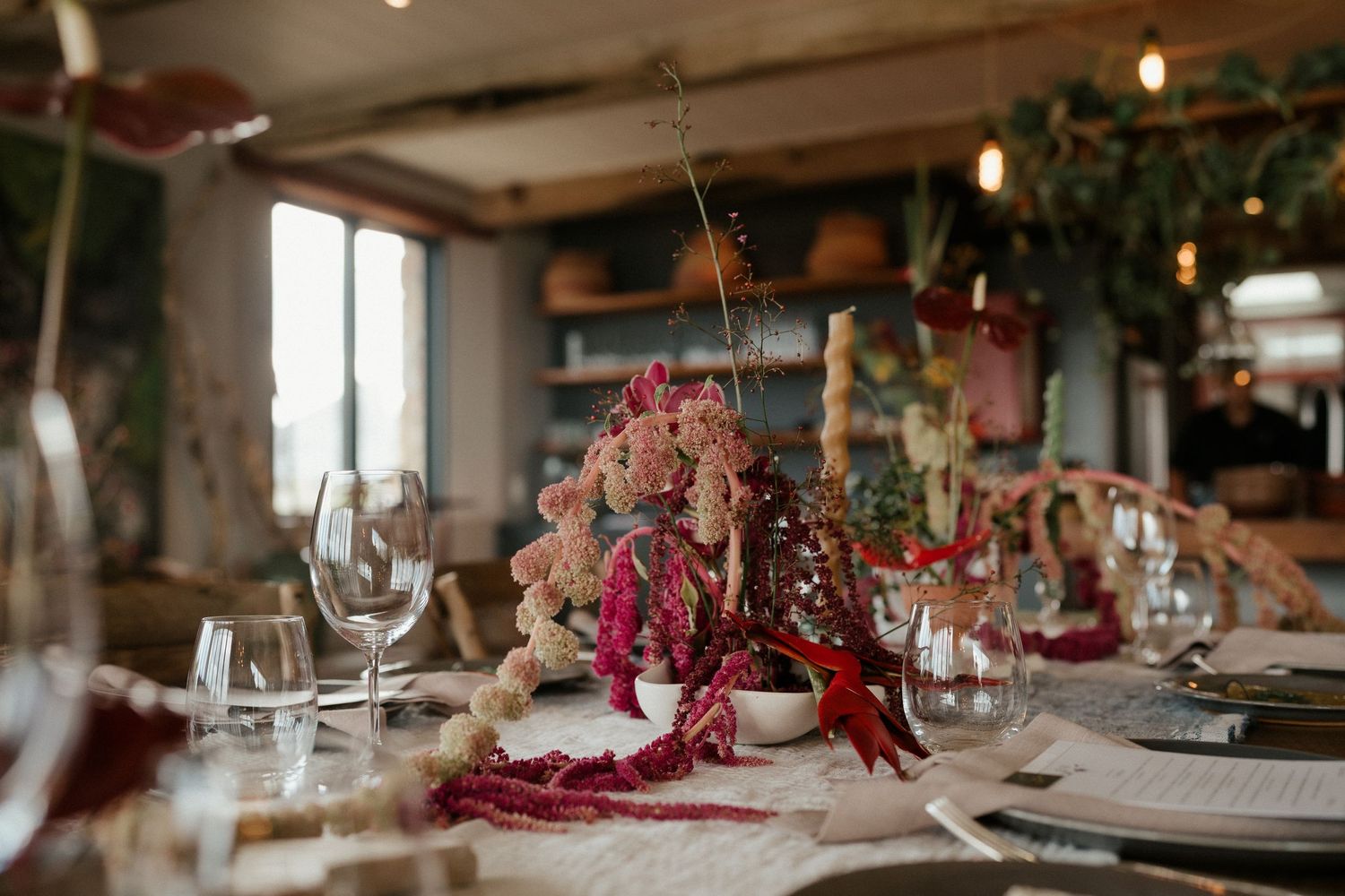 Romantic restaurant table setting with burgundy floral arrangements, wine glasses and moody lighting.