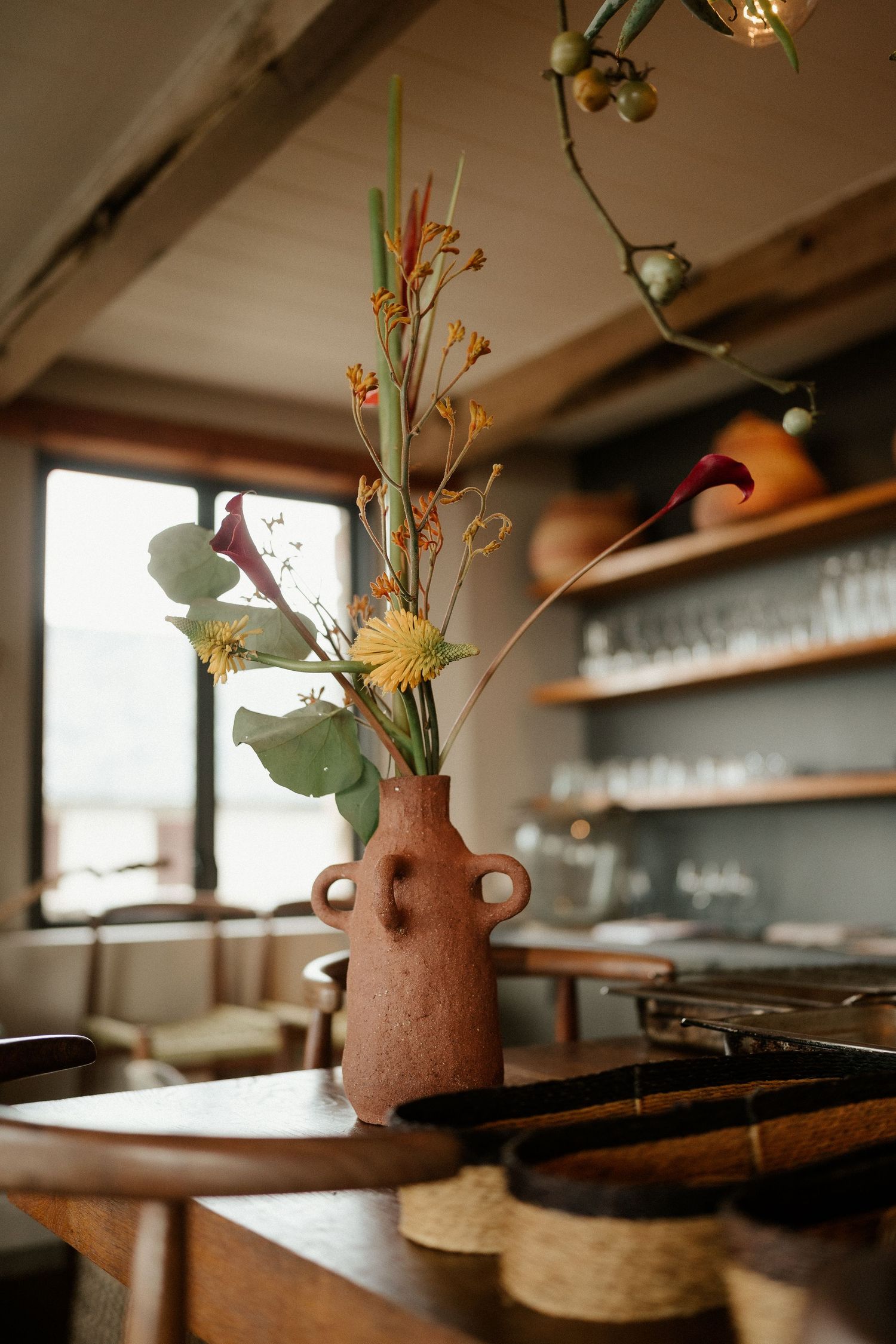 Terracotta vase with dried floral arrangement sits on wooden table in rustic restaurant setting.