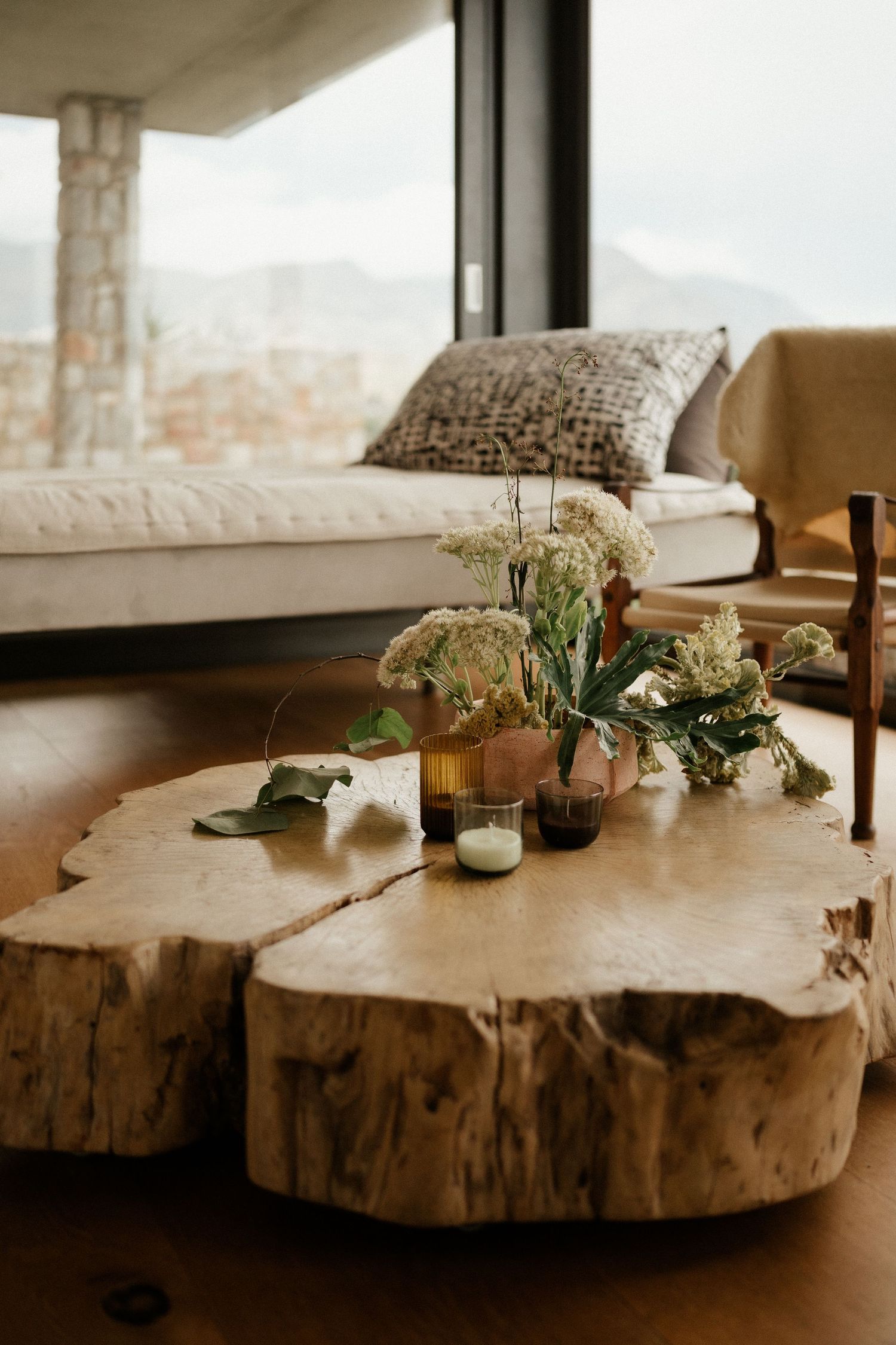A rustic live edge wood coffee table displays delicate flower arrangements and candles next to a cozy window seat.