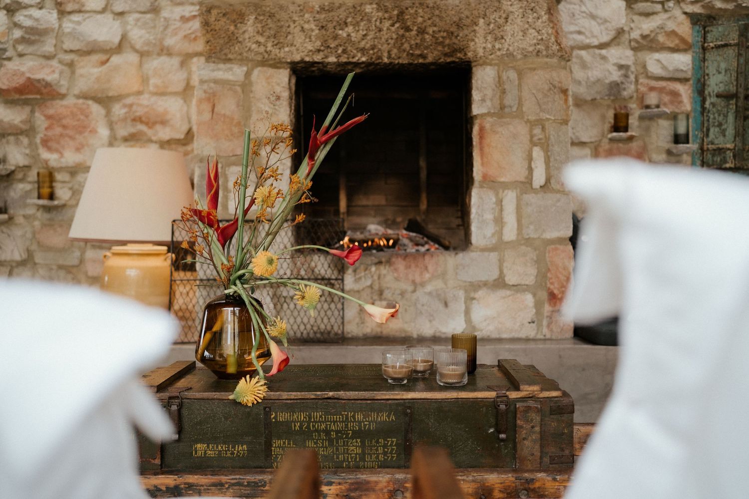 Rustic stone fireplace with decorative floral arrangement and vintage trunk creating a cozy wedding venue setting.
