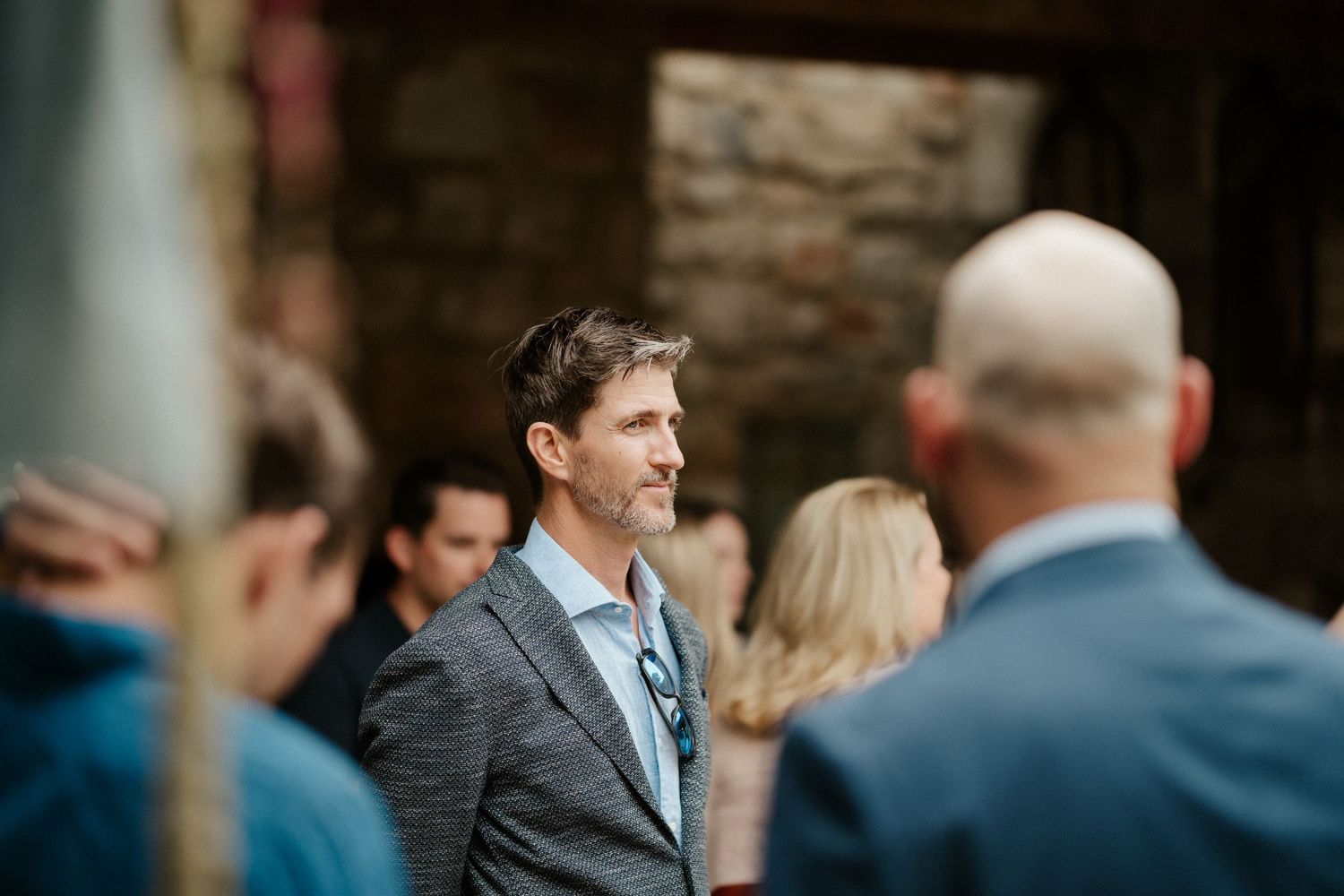Wedding guests gather outdoors with one person in a grey suit and blue tie standing in focus.