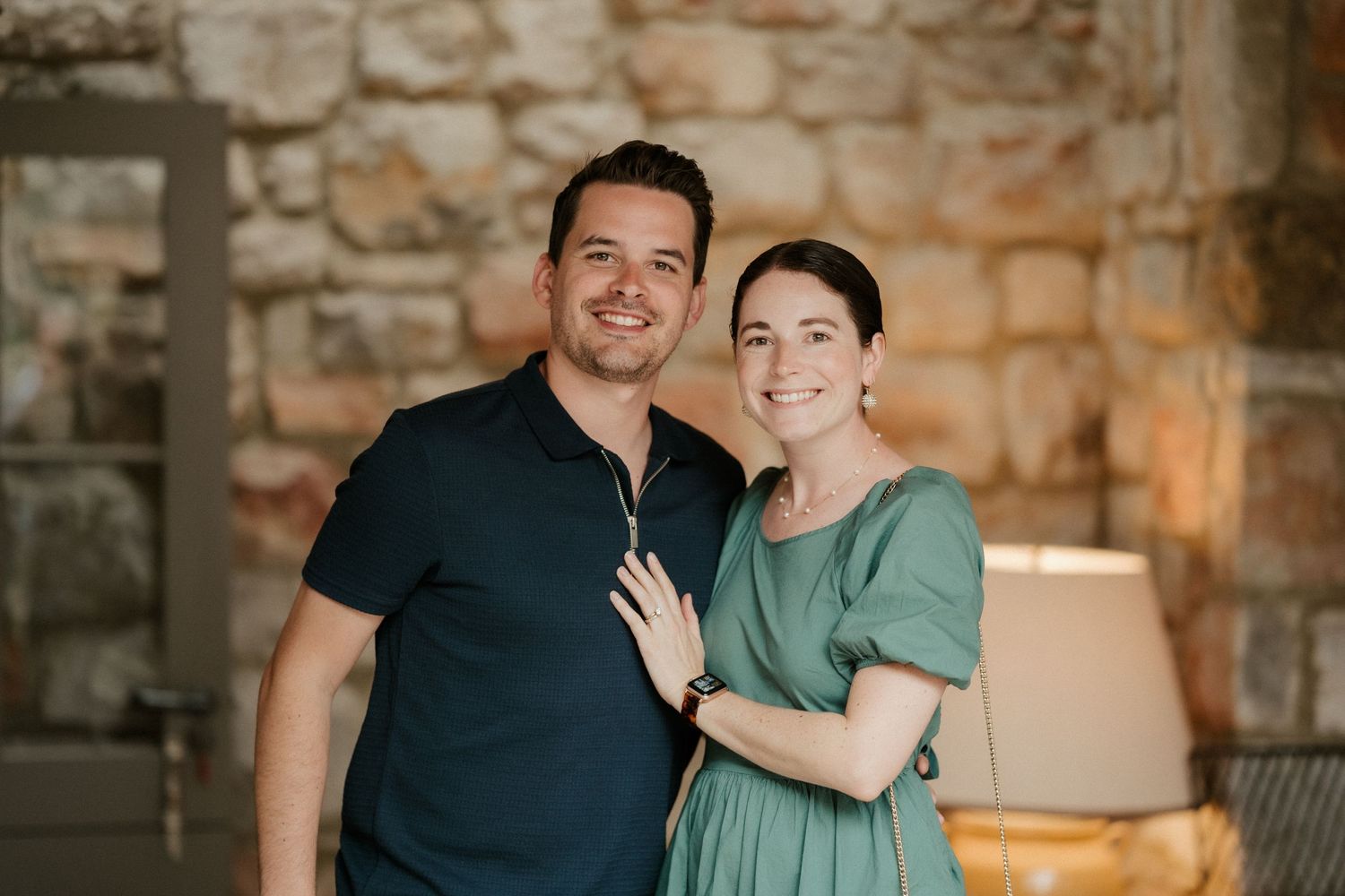 A couple stands together smiling in front of a stone wall with warm lighting.