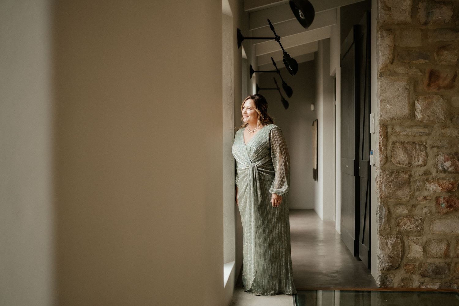 Someone stands in a long sage green dress by a window in a rustic stone hallway with natural lighting.