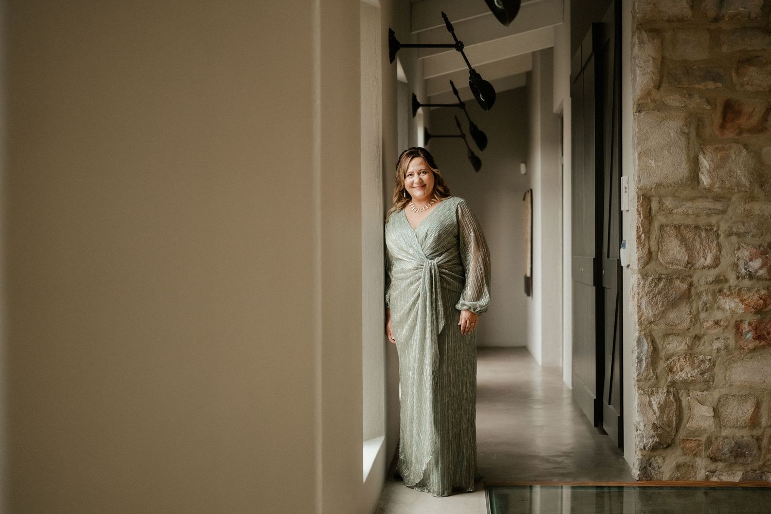 A person stands in an elegant sage green dress in a modern hallway with exposed stone walls.