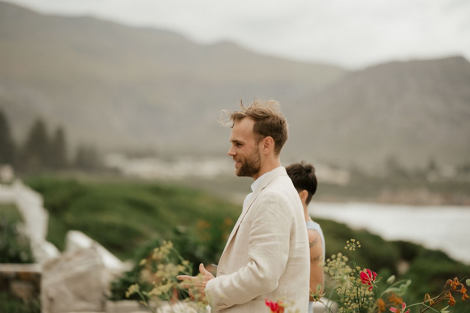 A couple stands against a scenic coastal backdrop with mountains and flowers.