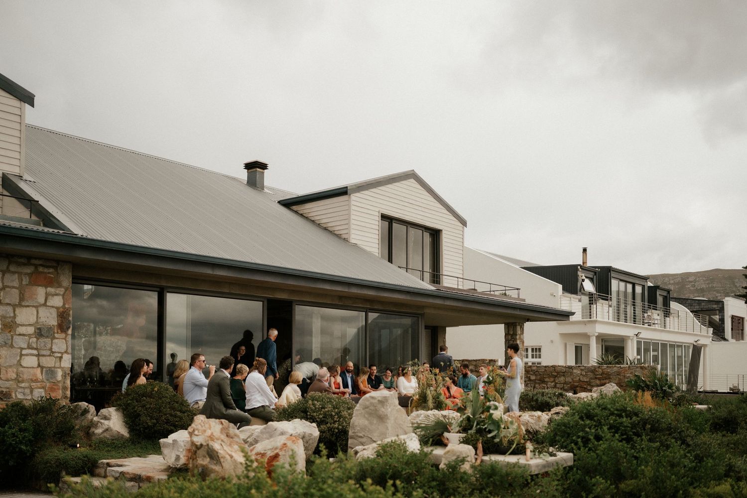 Contemporary stone house exterior with gathered guests on the patio under cloudy skies.