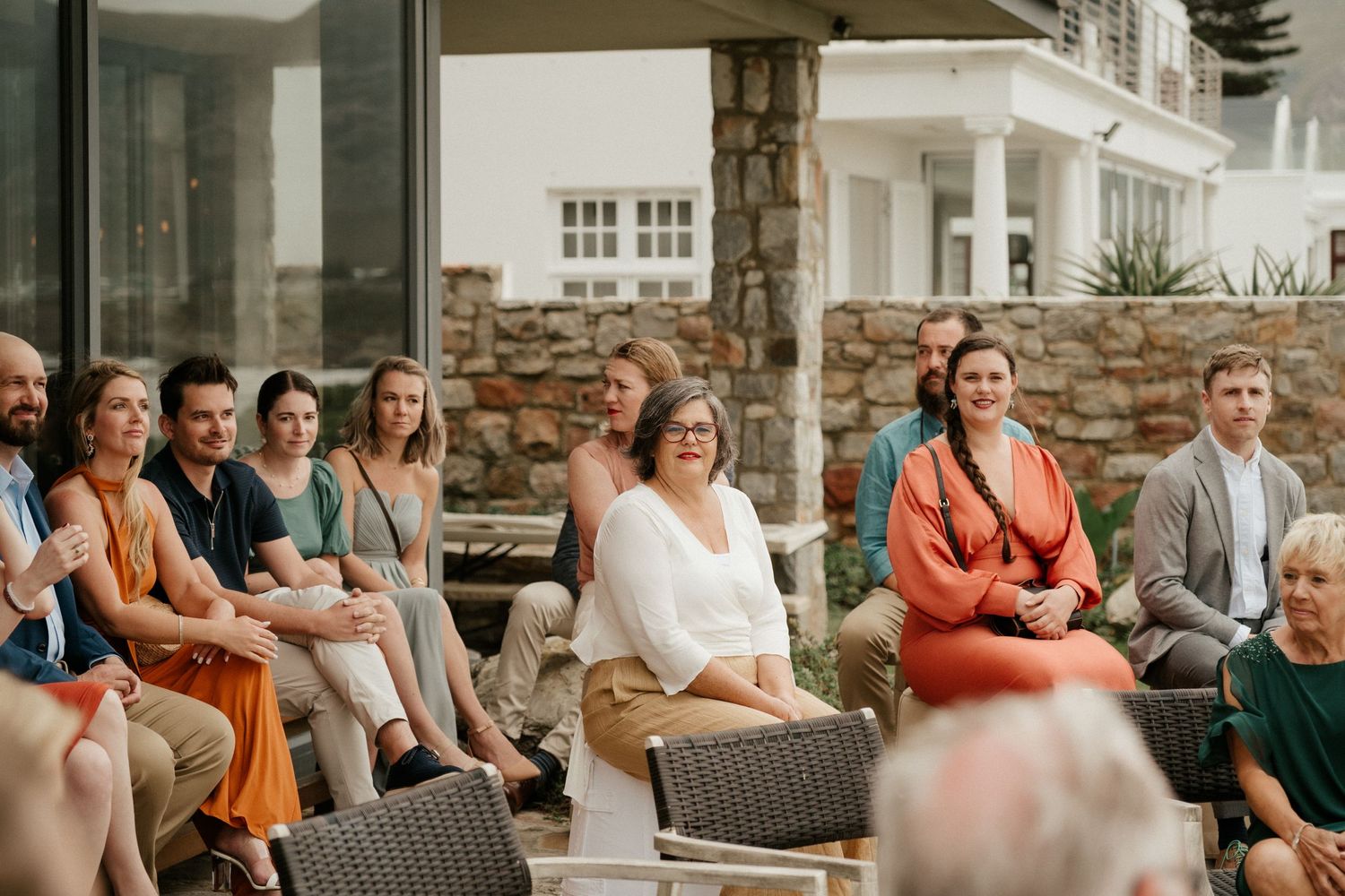 Wedding guests sit attentively on outdoor chairs during an elegant ceremony with stone architecture backdrop.