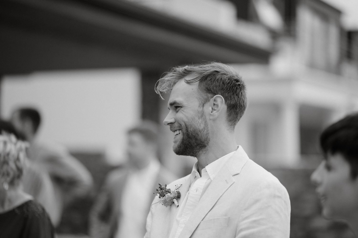 A groom in a white suit jacket smiles while attending an outdoor wedding celebration.