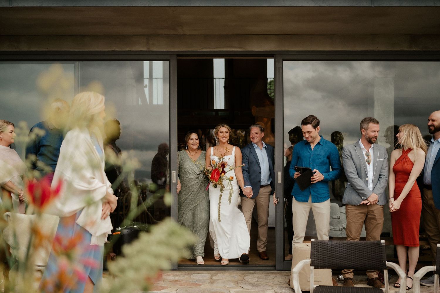 A wedding party gathers at a rustic doorway entrance with pastel floral decorations in the foreground.