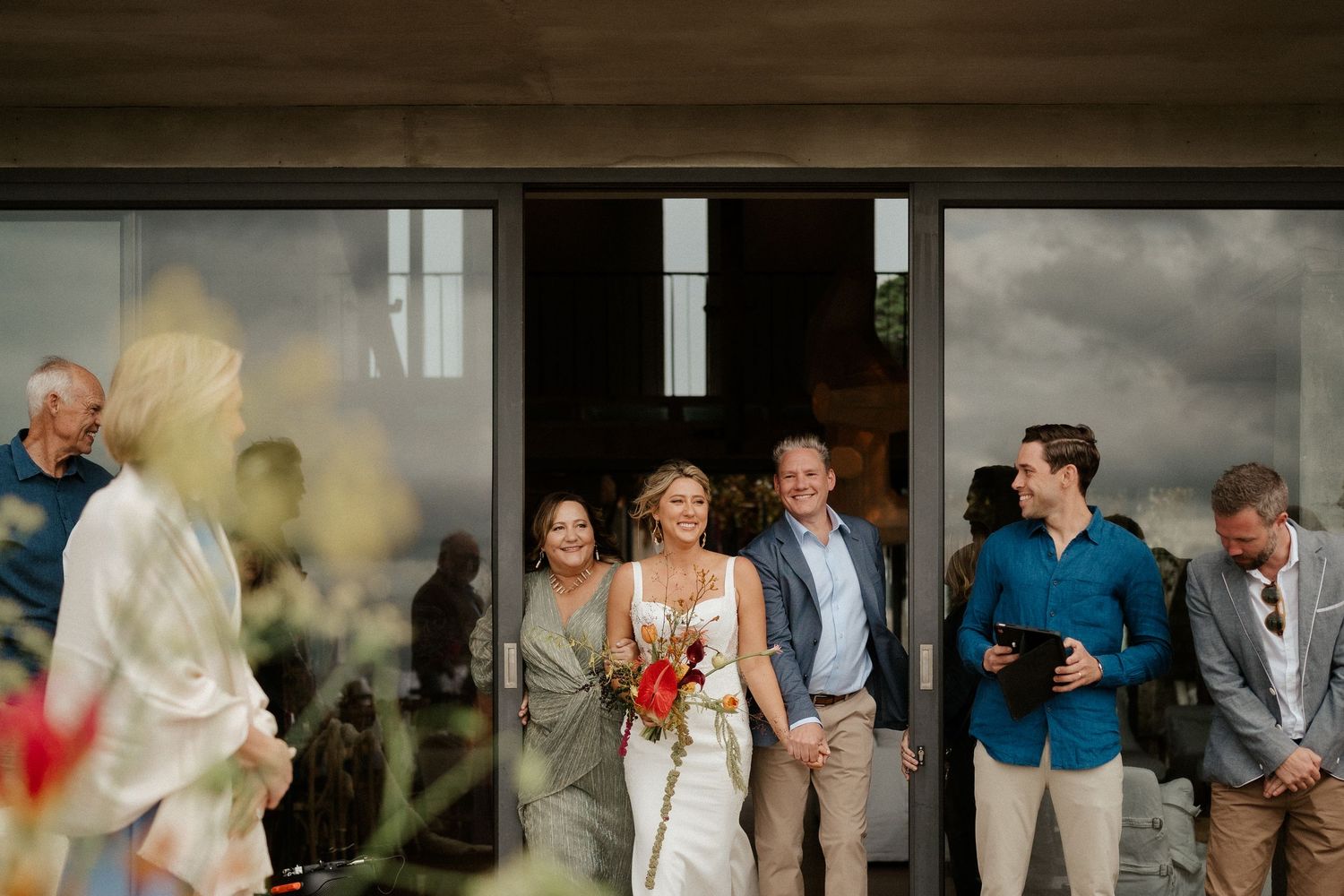 Wedding guests gather by large glass doors at an outdoor venue on an overcast day.