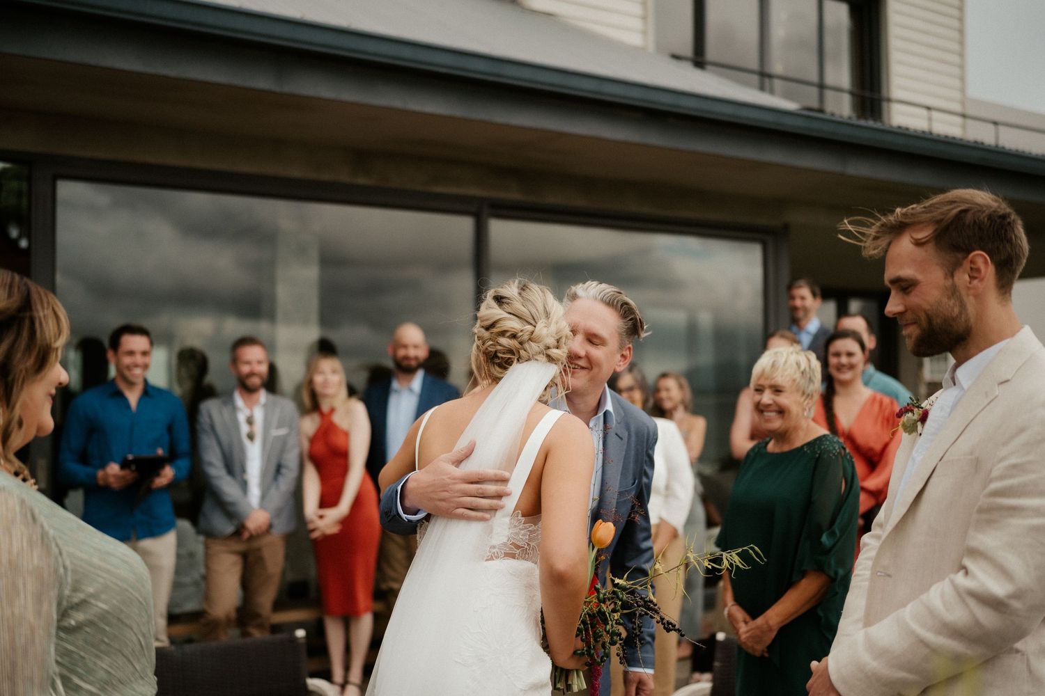 A wedding celebration moment with a couple embracing surrounded by guests during an outdoor daytime ceremony.