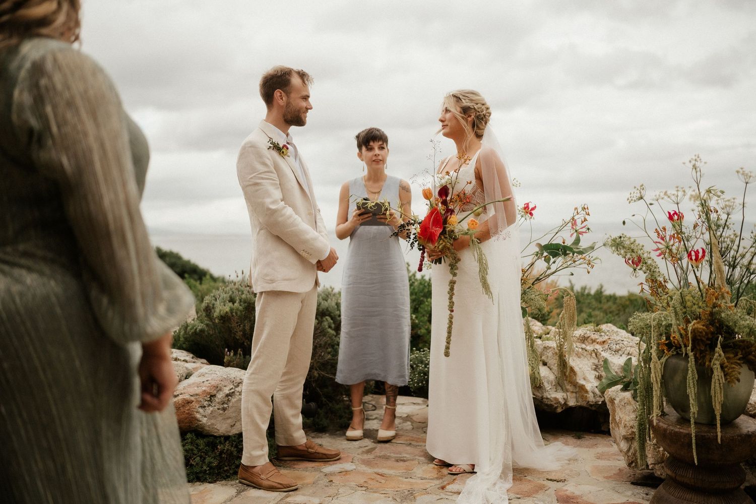An outdoor wedding ceremony takes place against a backdrop of mountains and cloudy skies.