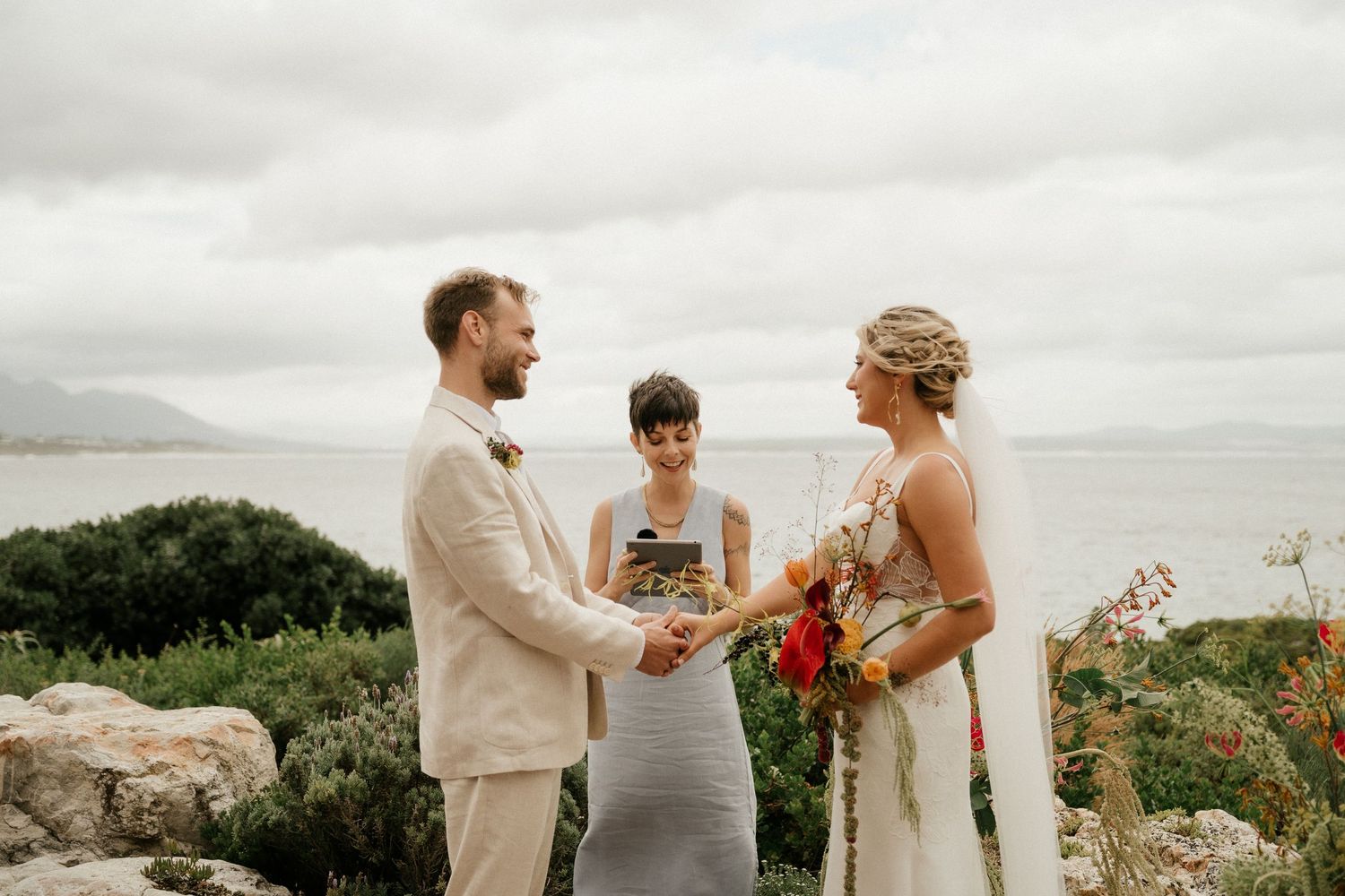 A wedding ceremony takes place on a scenic coastal clifftop with mountains and ocean in the background.