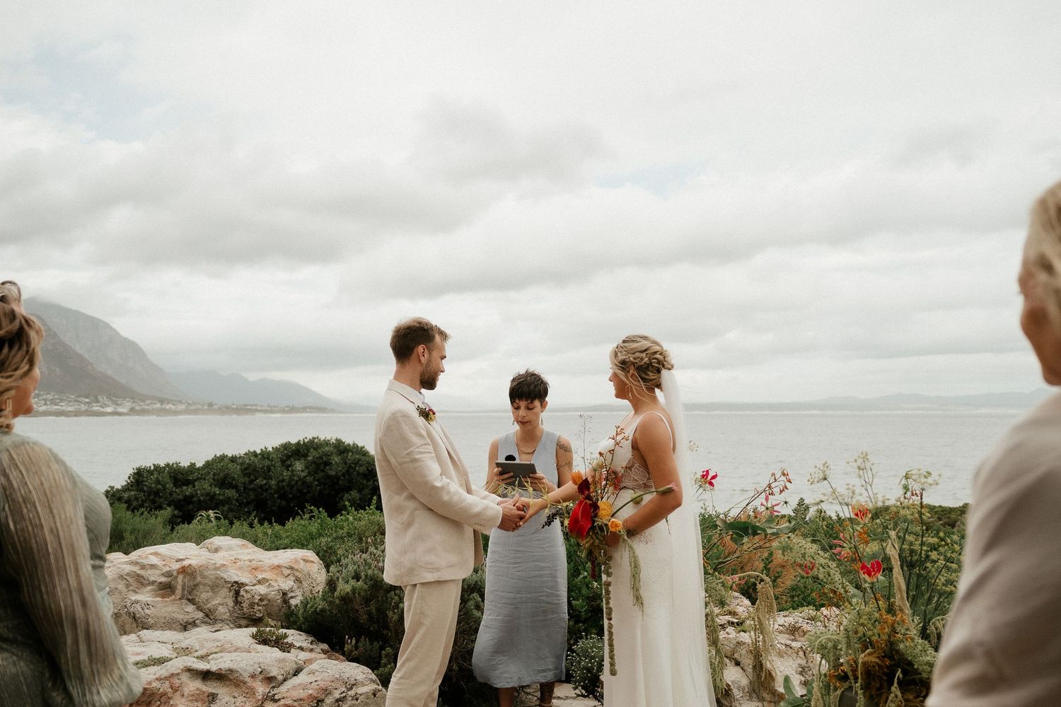 A romantic coastal wedding ceremony takes place on a rocky cliff overlooking the ocean under cloudy skies.