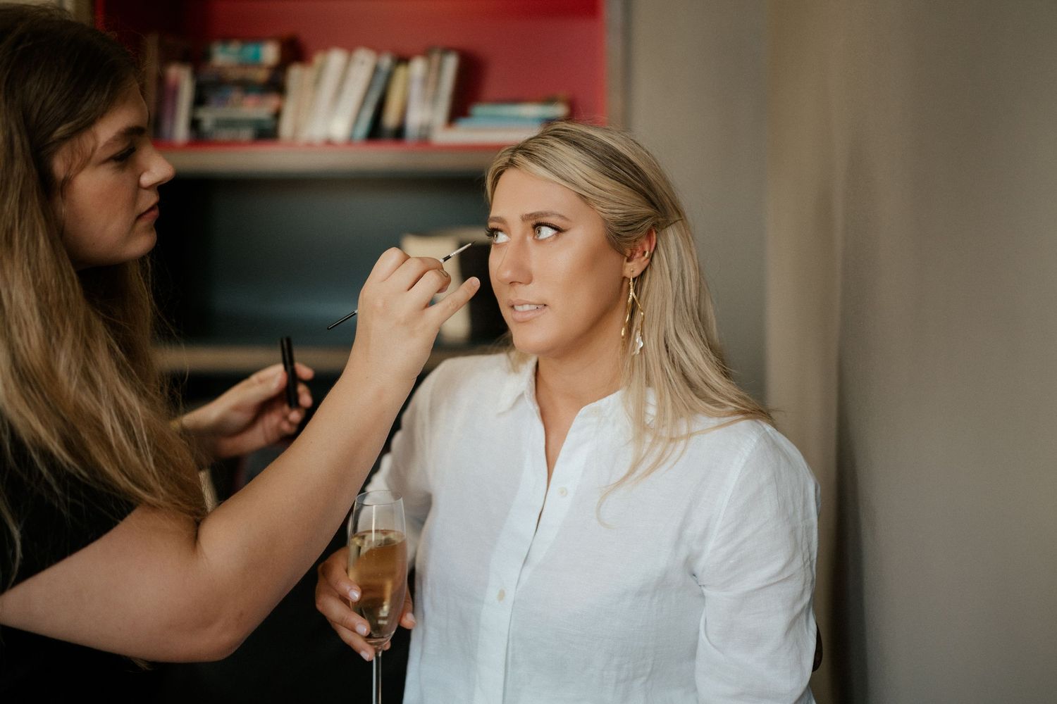 A bride gets her makeup professionally applied while wearing a white button down shirt during wedding preparations.