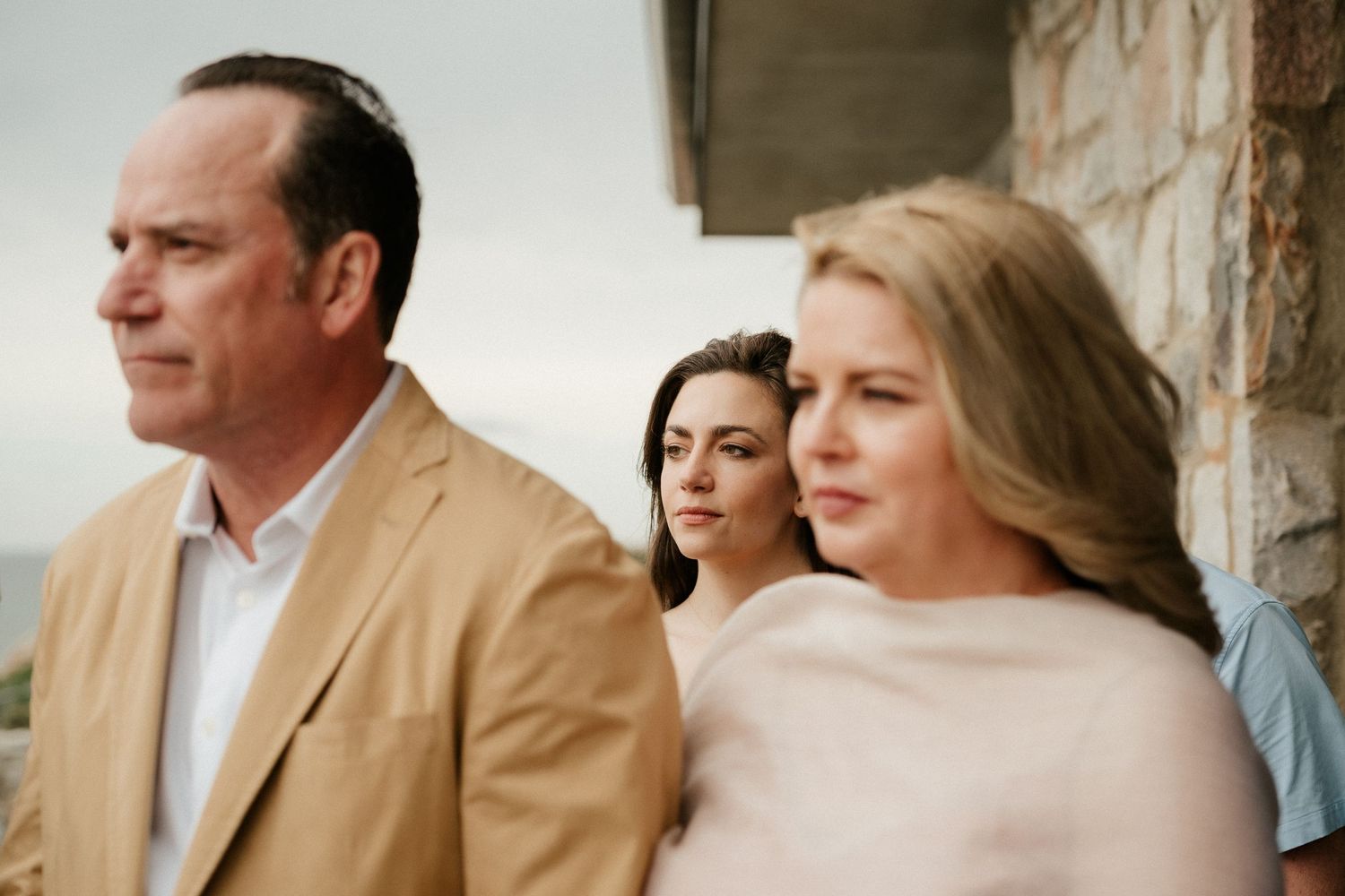 Three people stand together in front of a stone wall, wearing casual elegant attire on an overcast day.