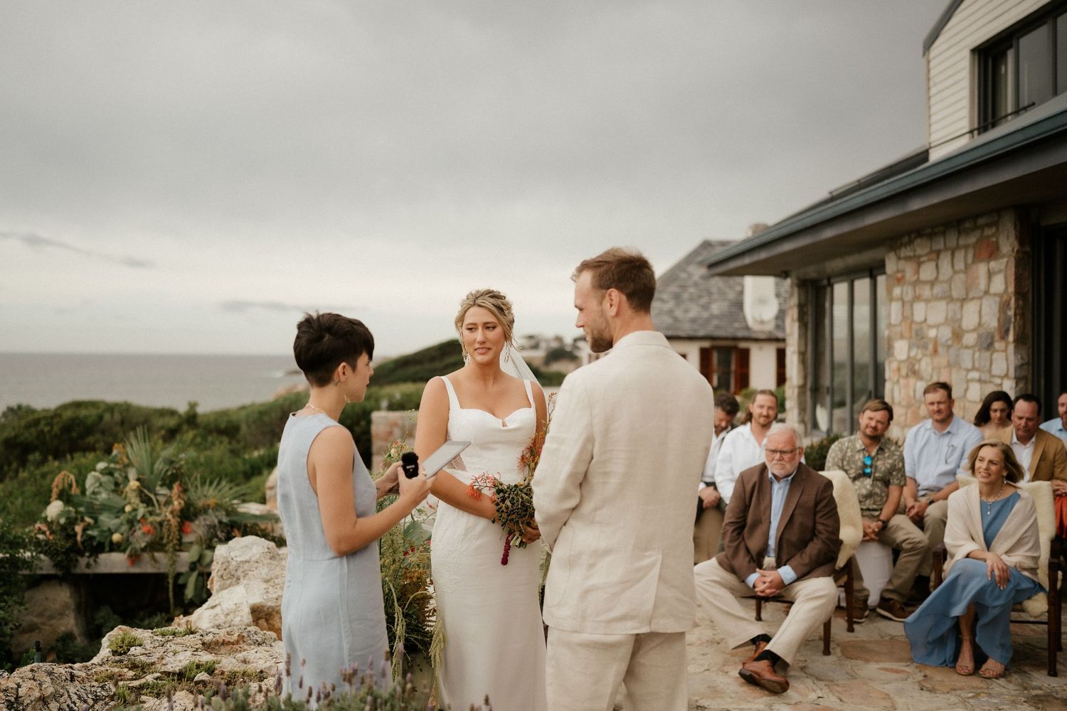 Intimate outdoor wedding ceremony taking place on a coastal cliff with guests seated on benches.