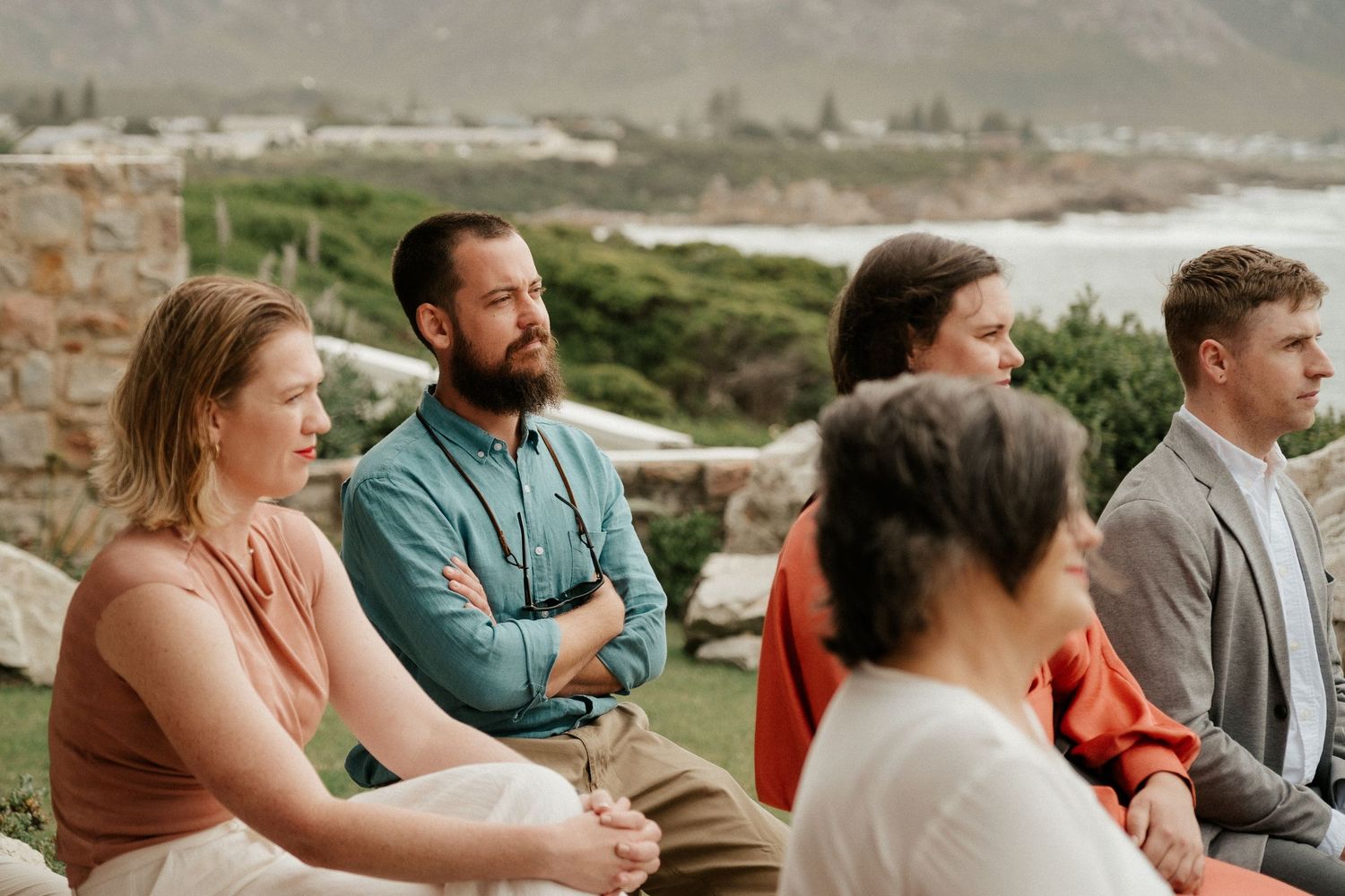 Wedding guests in casual formal attire seated outdoors watching ceremony with ocean views.