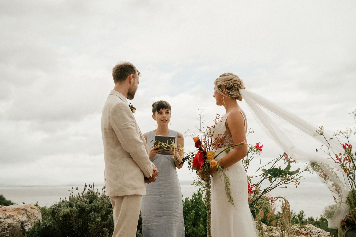 Outdoor wedding ceremony with flowing veil catching the breeze under cloudy skies.