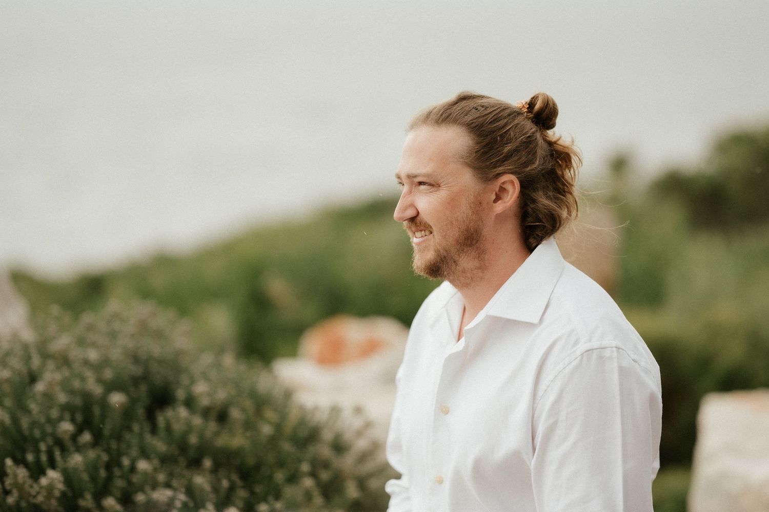 A person in a white button-down shirt smiles while looking to the side against a blurred green outdoor background.