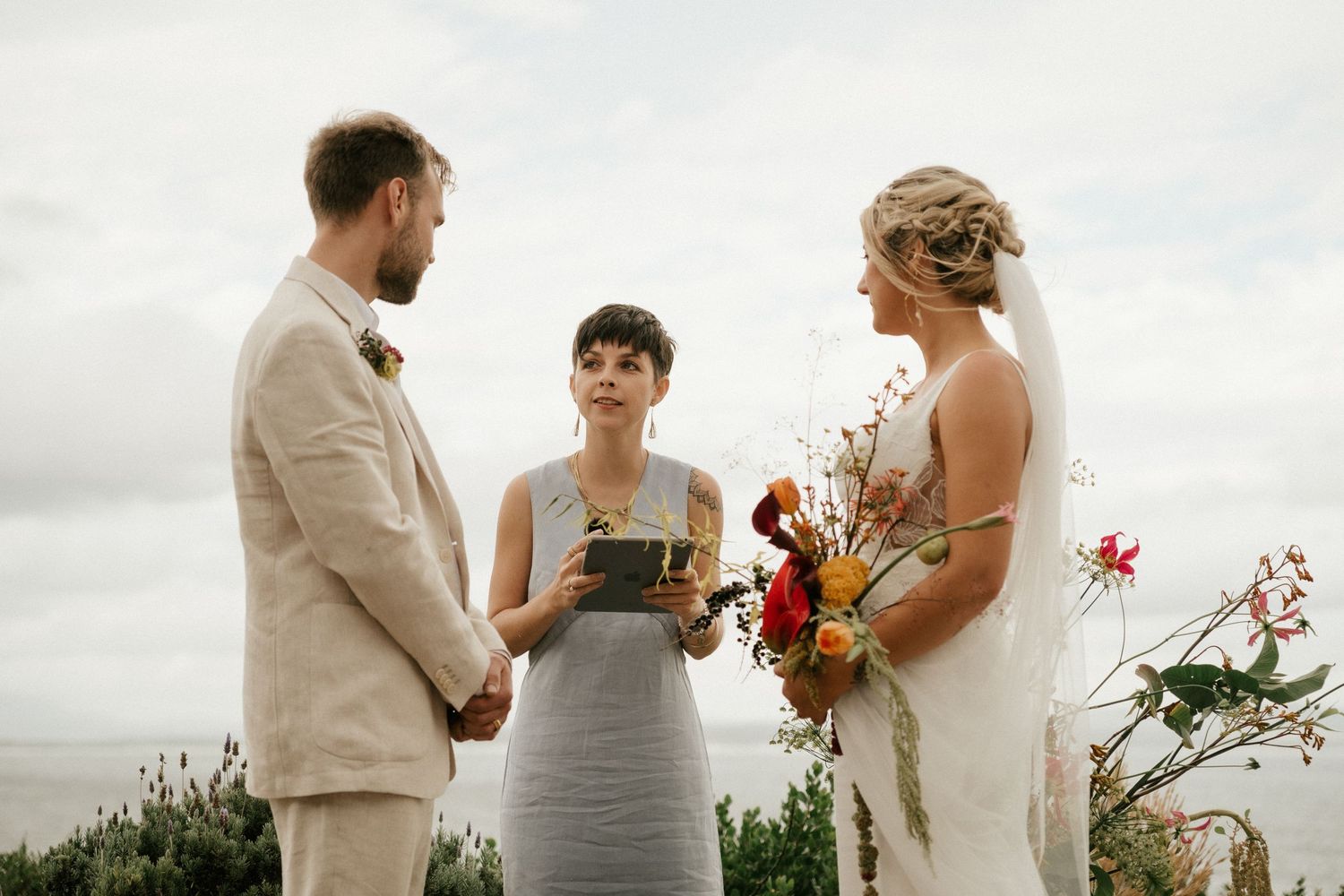 Wedding ceremony on a coastal backdrop with an officiant and couple exchanging vows with autumn-colored bouquet.