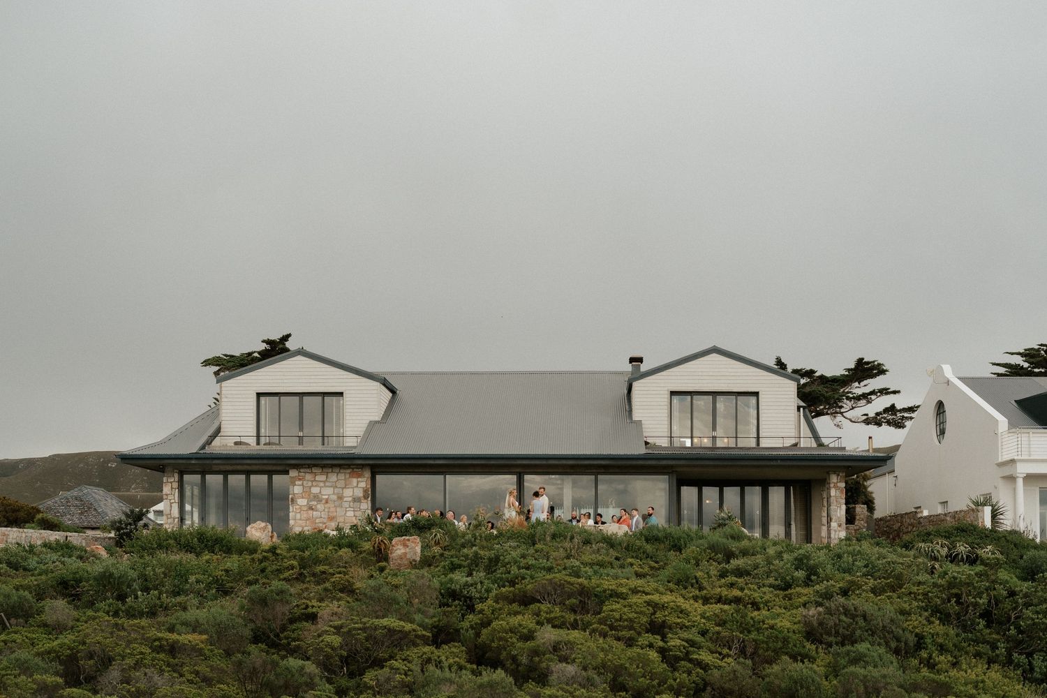 Modern coastal house with stone facade and gray roof nestled among seaside vegetation.