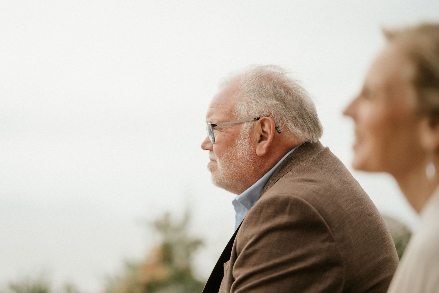 An elderly person in a brown jacket gazes thoughtfully into the distance on an overcast day.