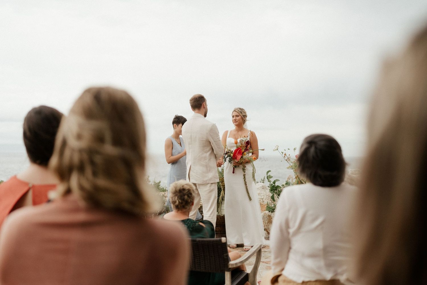 An intimate outdoor wedding ceremony takes place with guests watching by the water on a bright day.
