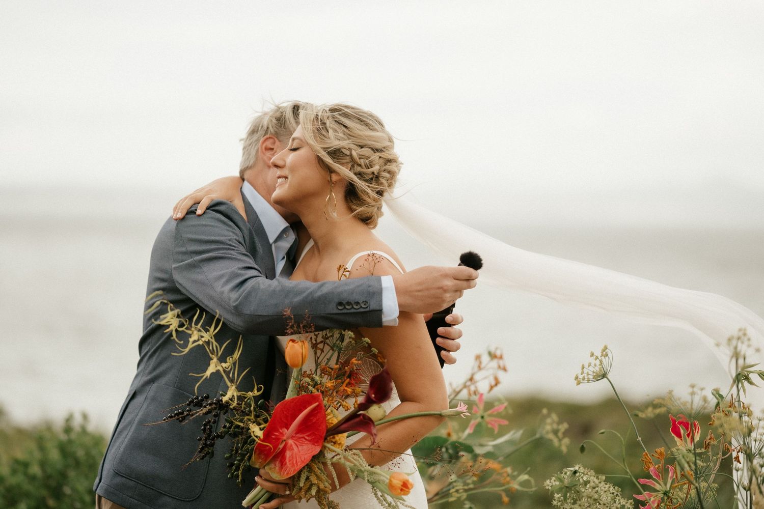 Intimate wedding moment captured against overcast sky with red floral bouquet.