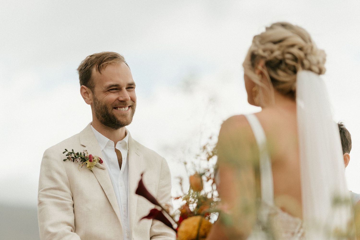 A beaming groom in a cream suit with a boutonniere shares a joyful moment during an outdoor wedding ceremony.