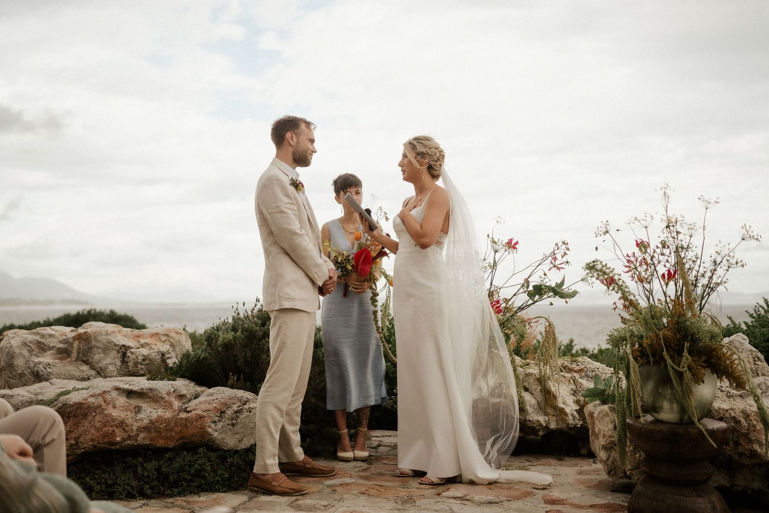 A wedding ceremony takes place on a rocky outcrop overlooking a scenic landscape with potted flowers as decor.