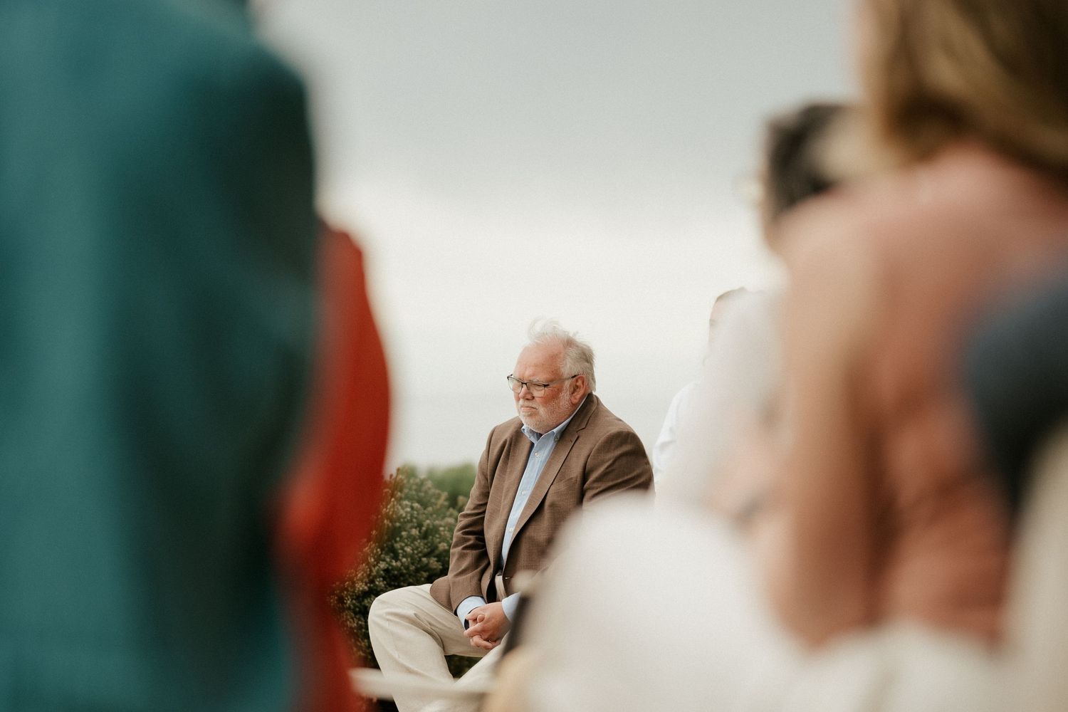 An older person sits outdoors during what appears to be a wedding ceremony.