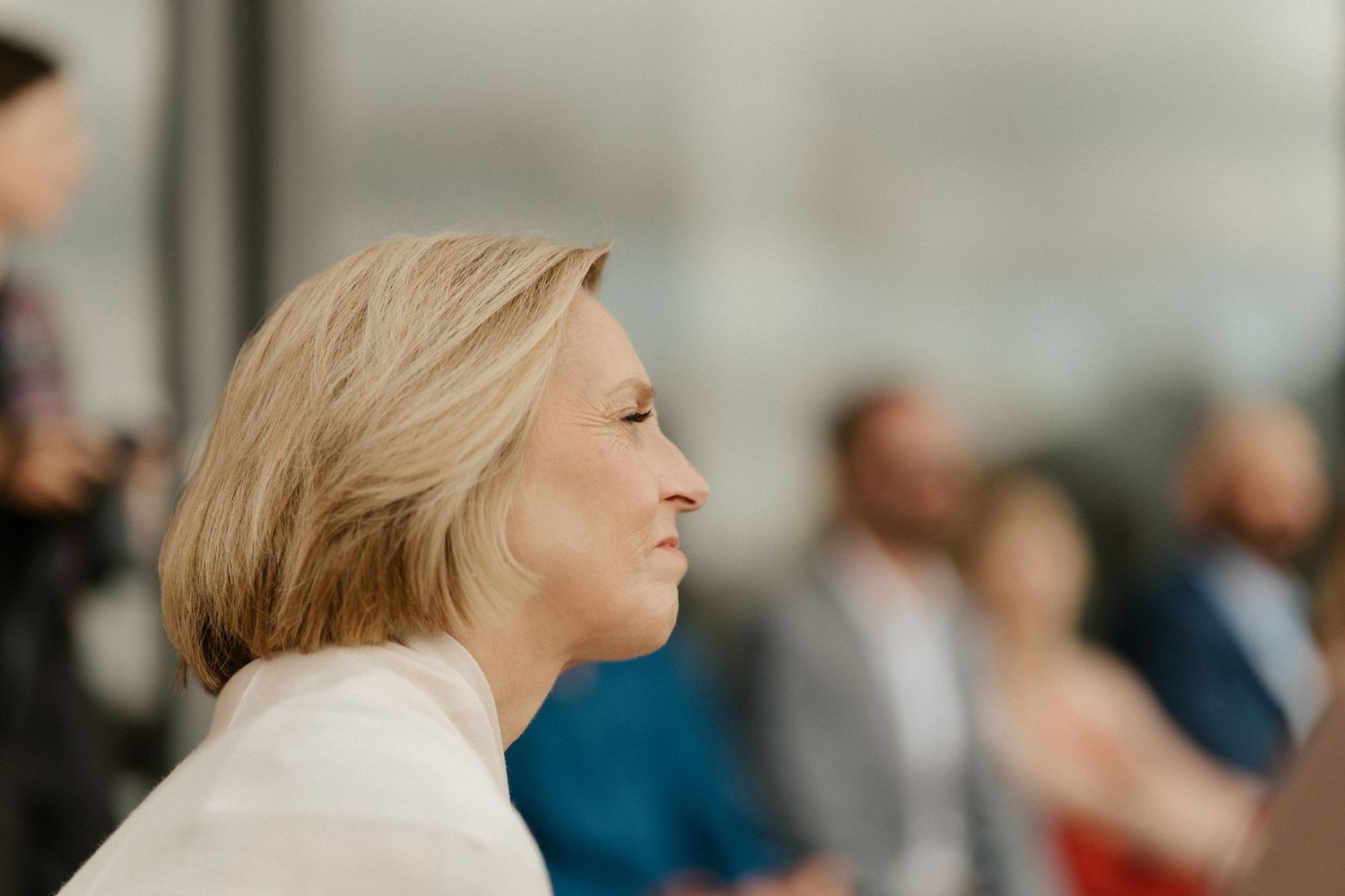 A person in a white blazer looks attentively to the side during an indoor event.
