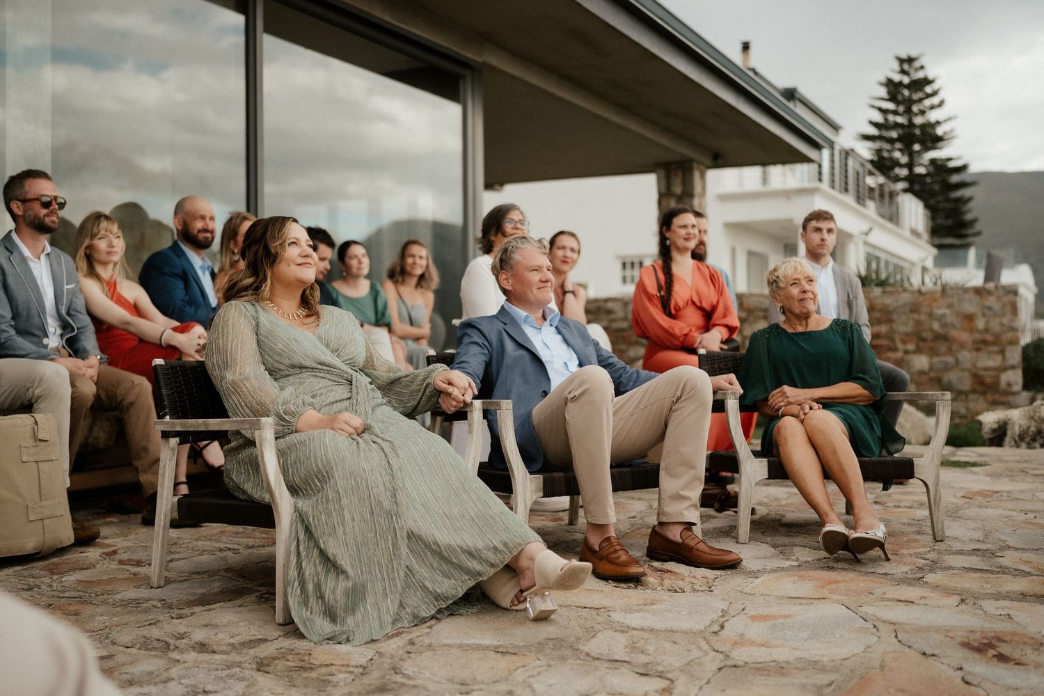 A group of guests sit on a stone patio watching an outdoor wedding ceremony with mountains in the background.