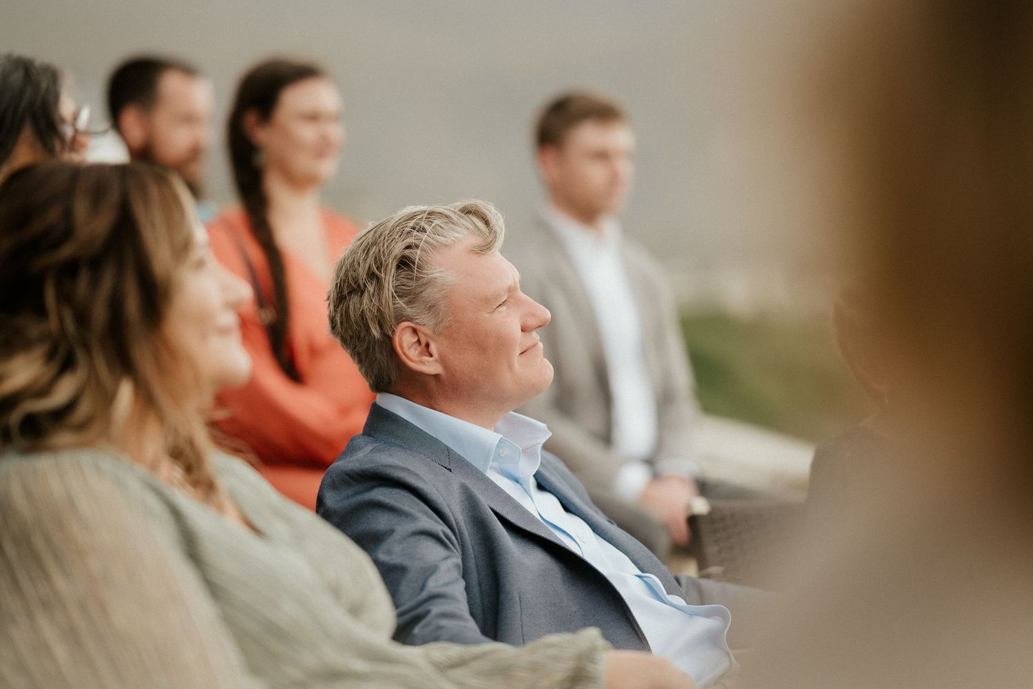 Wedding guests smile and laugh during an outdoor ceremony on a sunny afternoon.