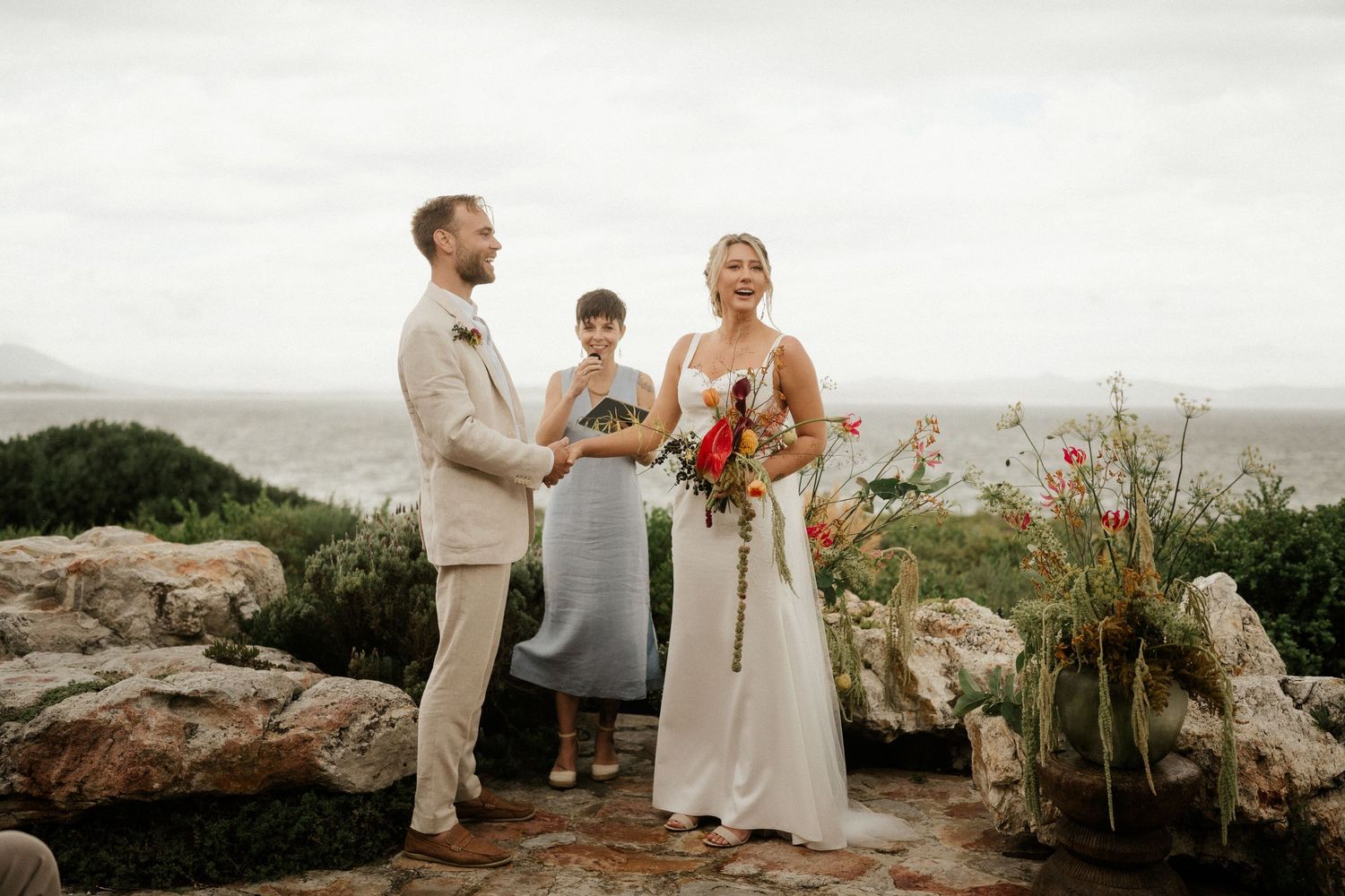 A couple stands on rocky cliff overlooking ocean during their intimate wedding ceremony.
