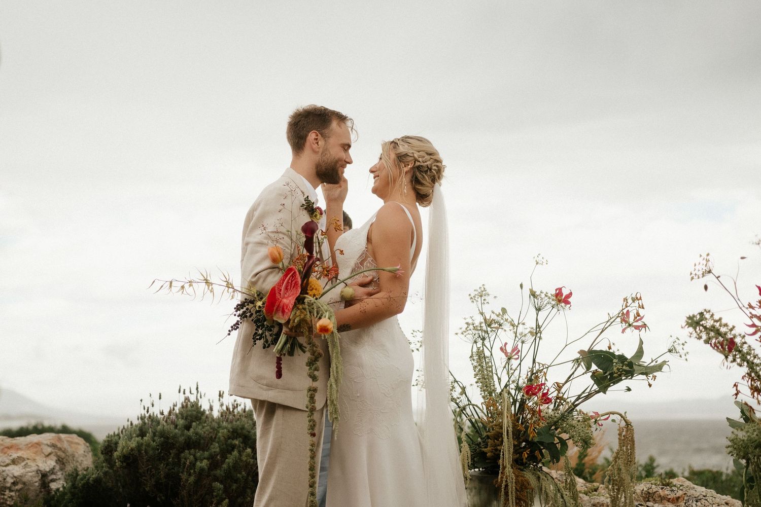 A romantic coastal wedding moment with red floral bouquet accents against an overcast sky.