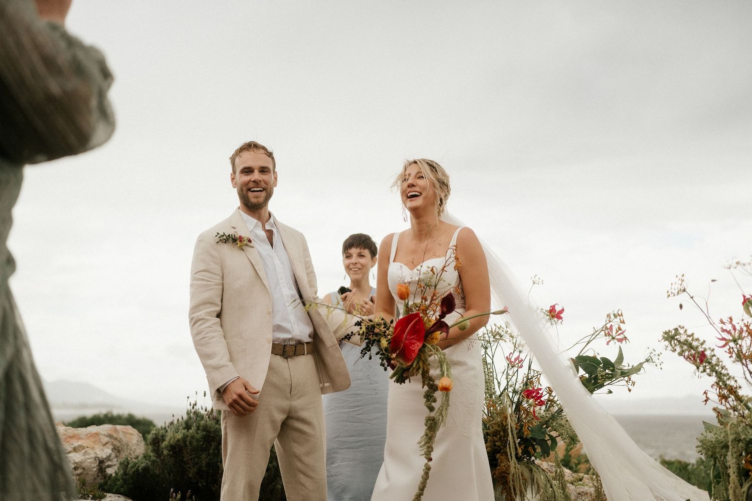 A couple shares a joyful moment during an outdoor wedding ceremony against an overcast sky.
