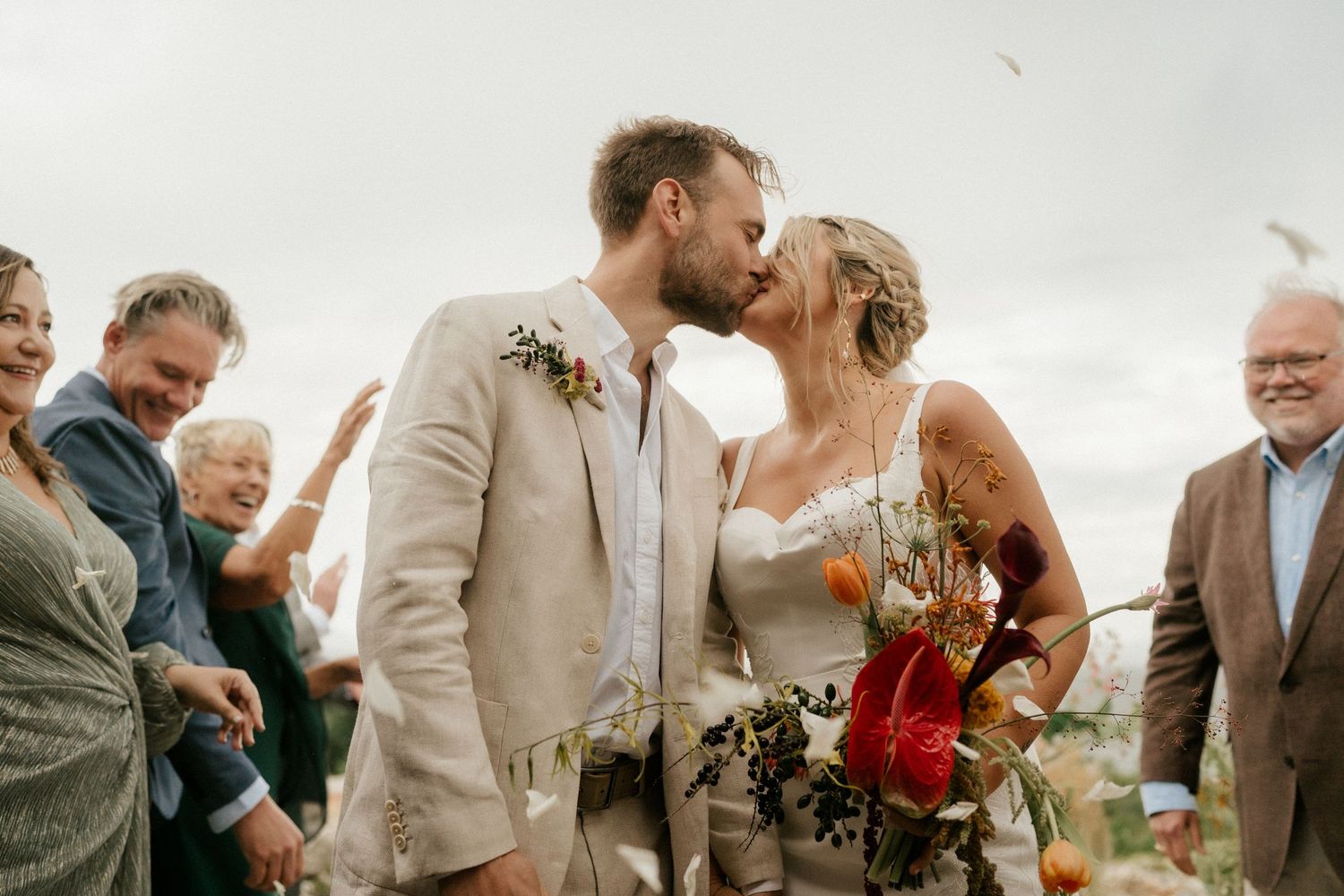 A couple shares their first kiss as newlyweds while guests celebrate with confetti in the background.