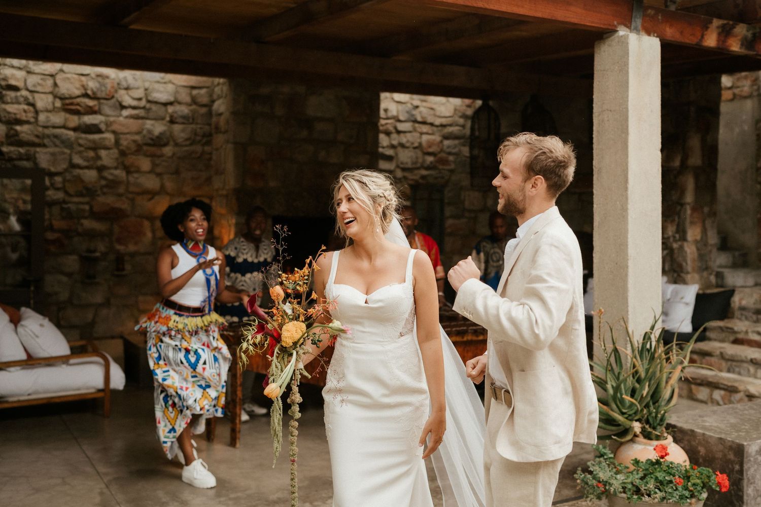 A couple in wedding attire celebrate on a stone patio while guests dance in the background.