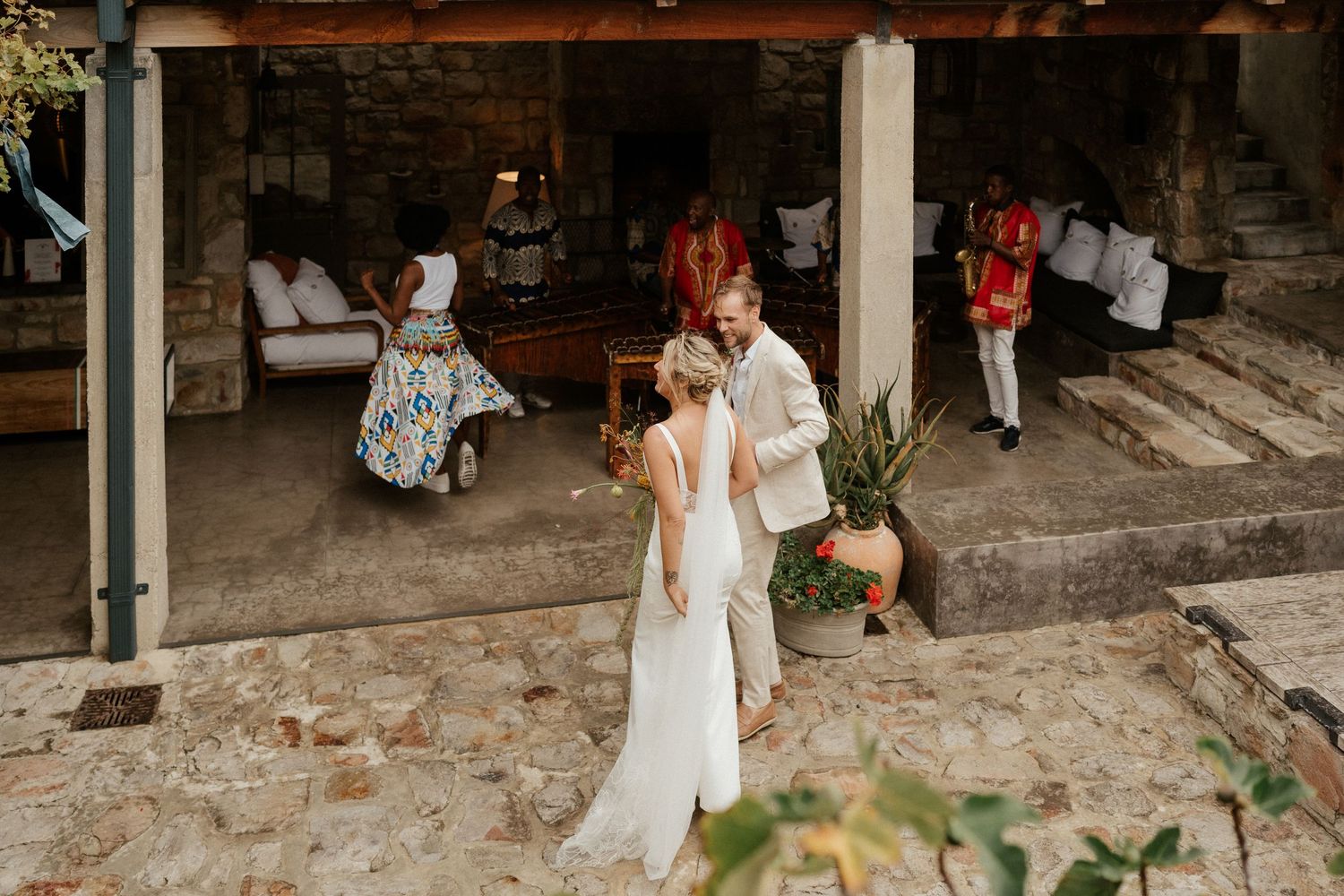 A wedding celebration scene at a rustic stone venue with guests gathering under an outdoor covered area.